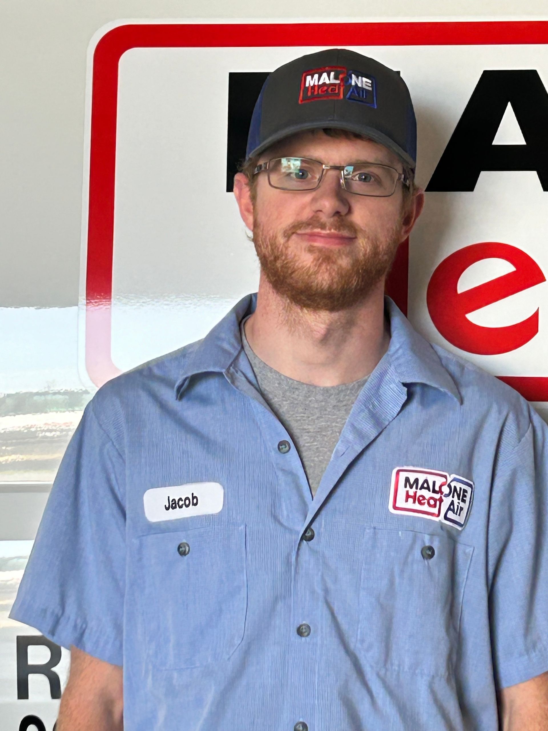 Man wearing a blue work shirt and cap, standing in front of a sign. He has a name tag that reads 