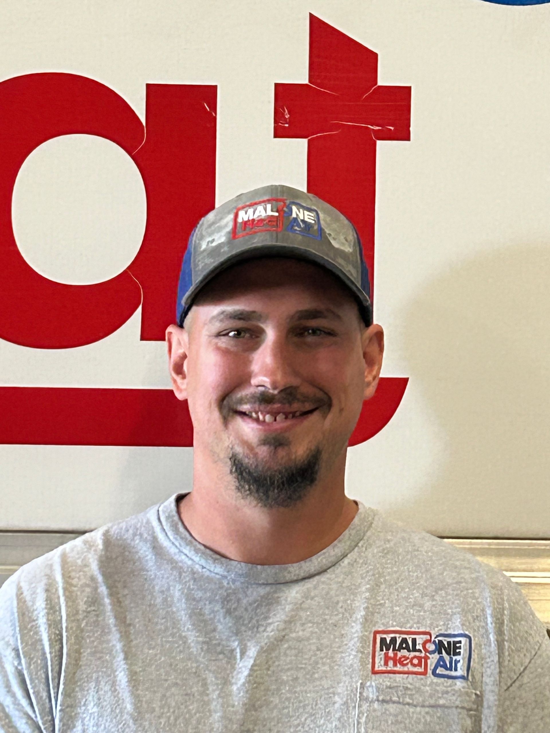 Man wearing a hat and gray shirt smiles in front of a sign.