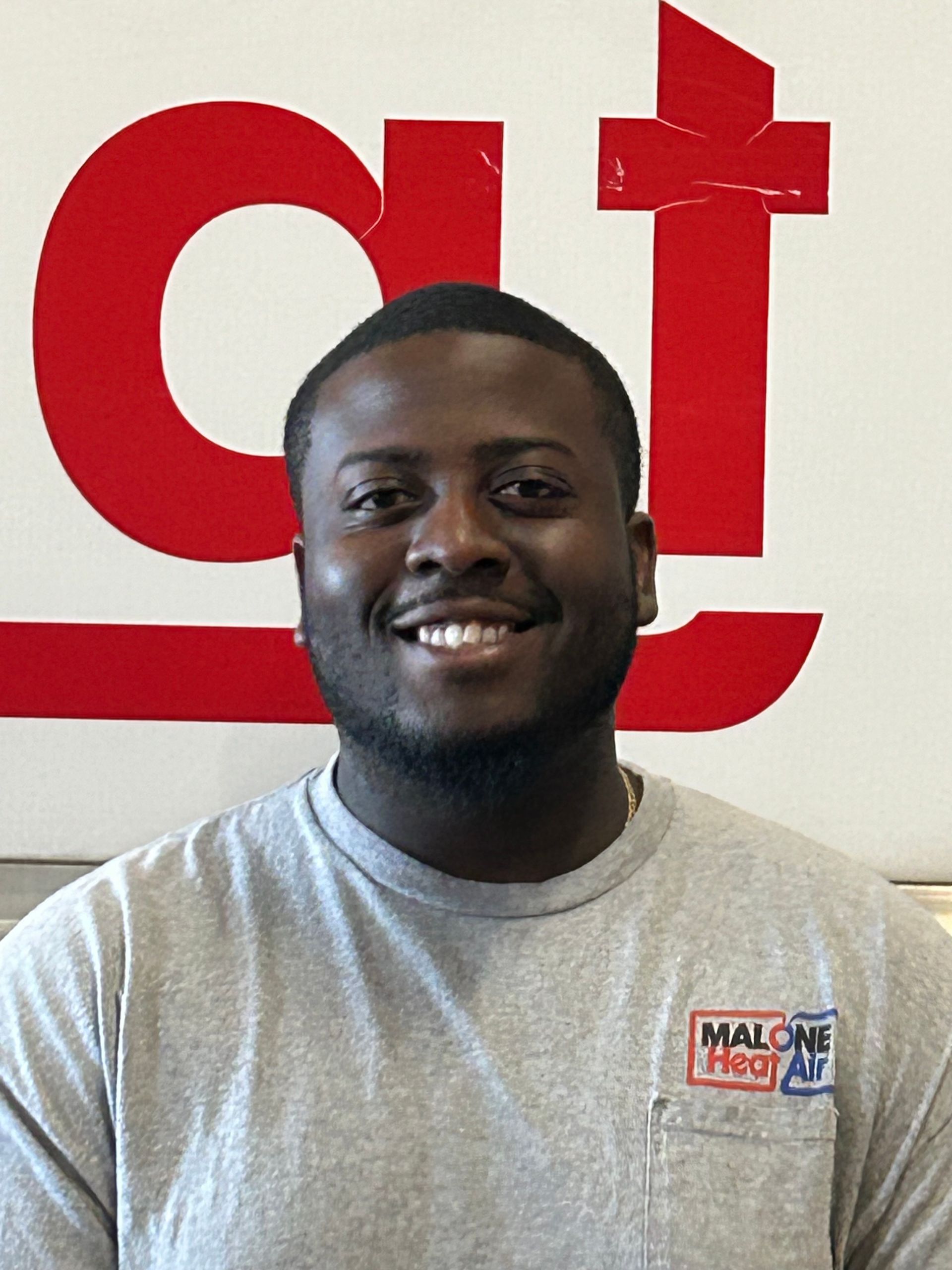 Man smiling in front of a red and white logo. He wears a gray t-shirt with a pocket.
