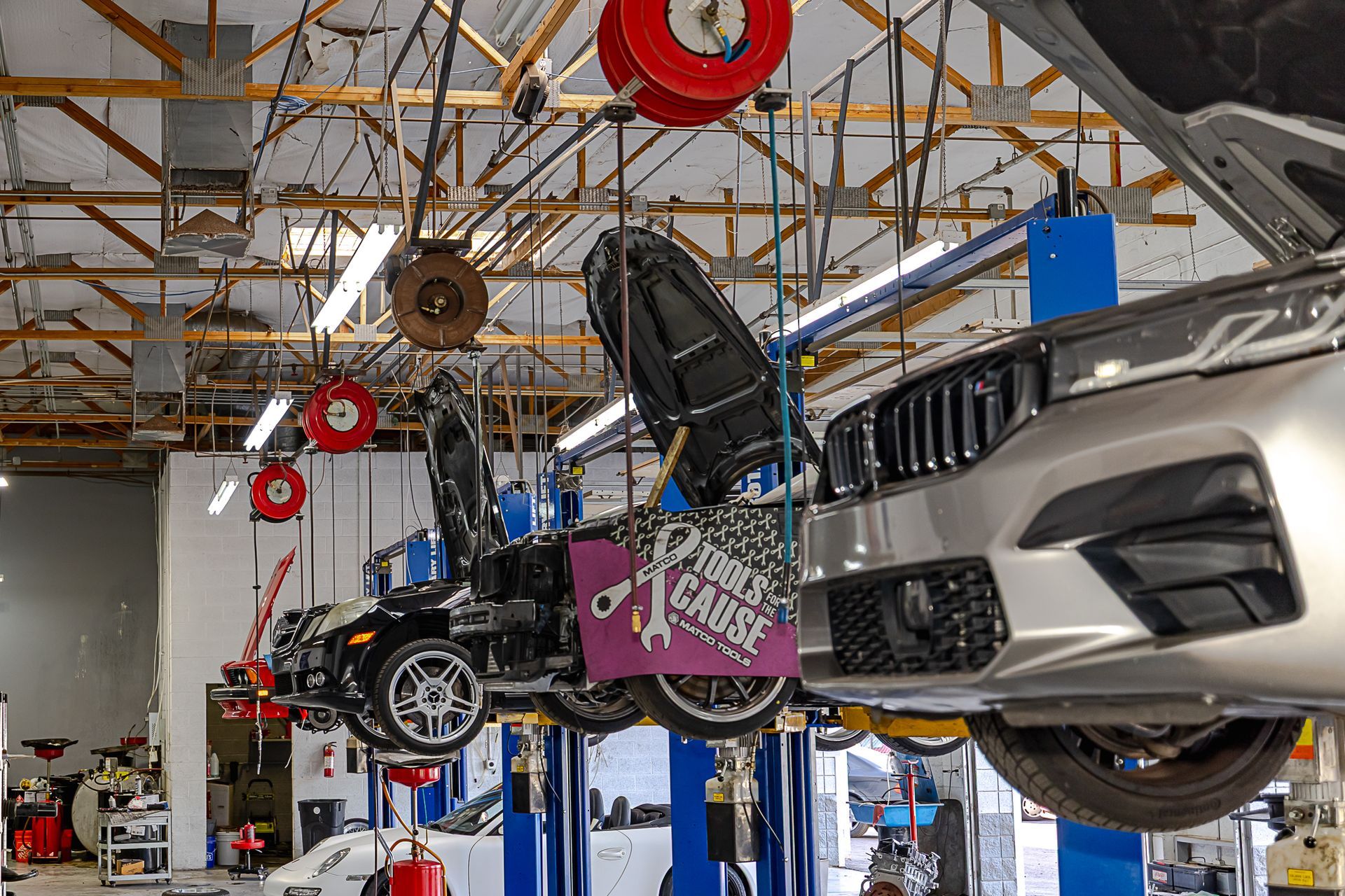 Cars on lifts inside auto repair shop; silver BMW and black Jeep with open hoods. Workshop has tools overhead.