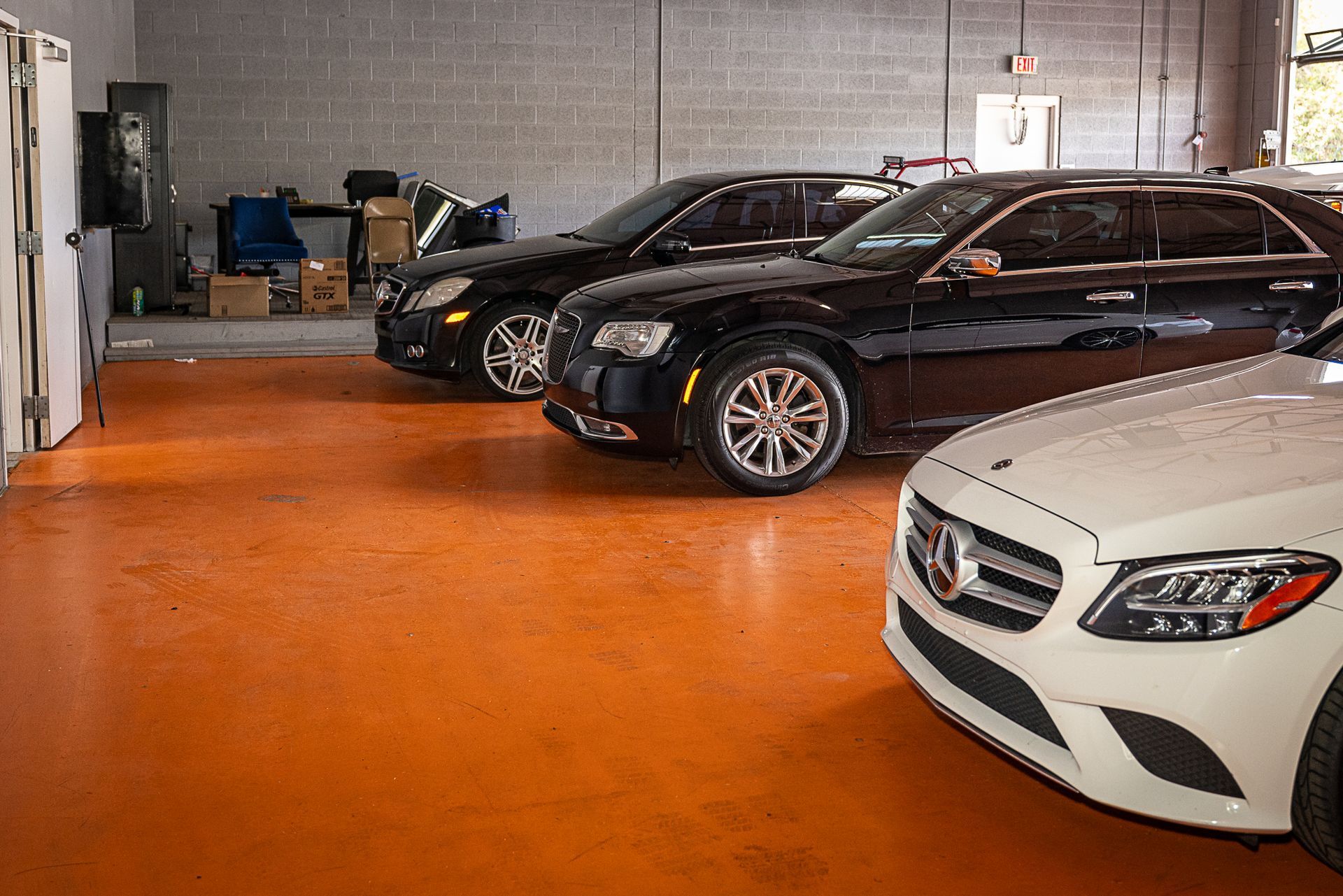 Three cars parked inside a garage. One white, two black, orange floor, and a brick wall.