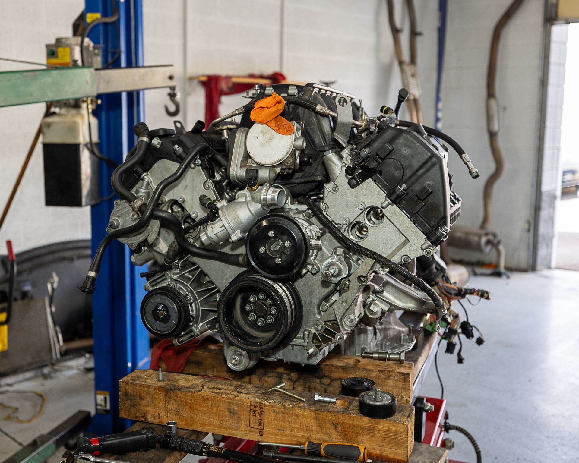 Car engine on a wooden workbench in a garage setting with various components and tools visible.