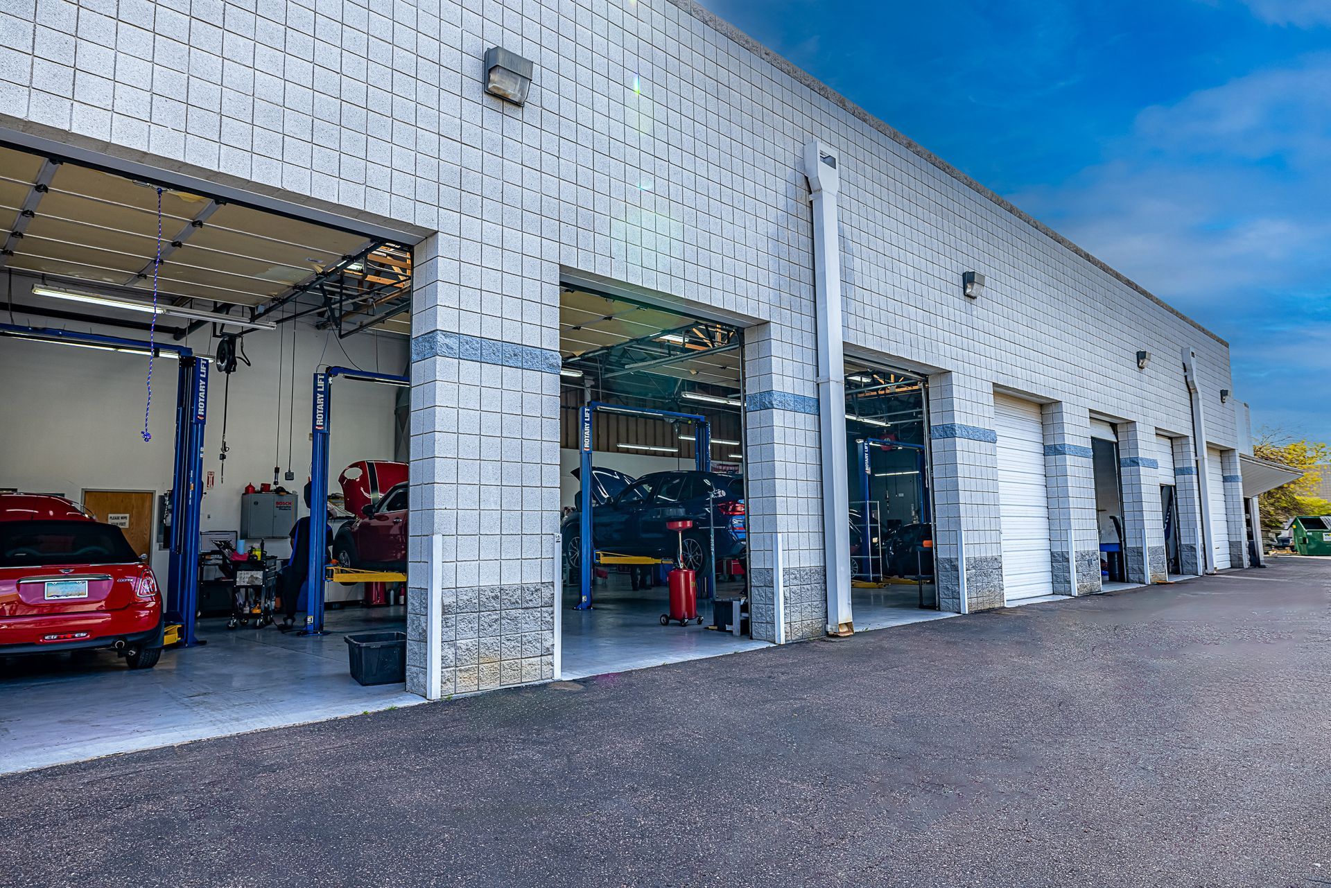 Exterior view of a car repair shop with several open bays, red car inside one bay, and blue sky.