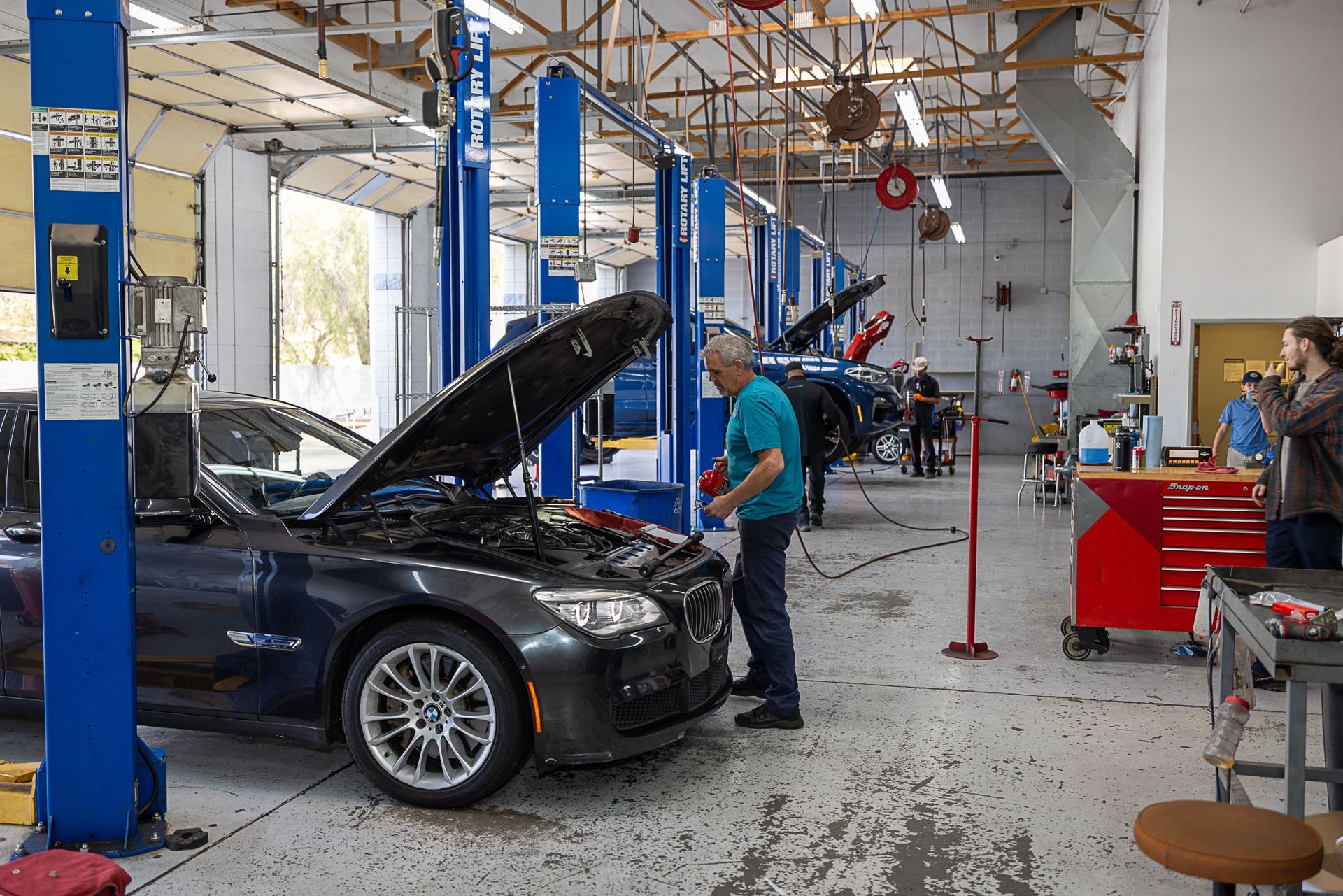 Man working on a car with the hood open in a garage, other cars in the background on lifts.