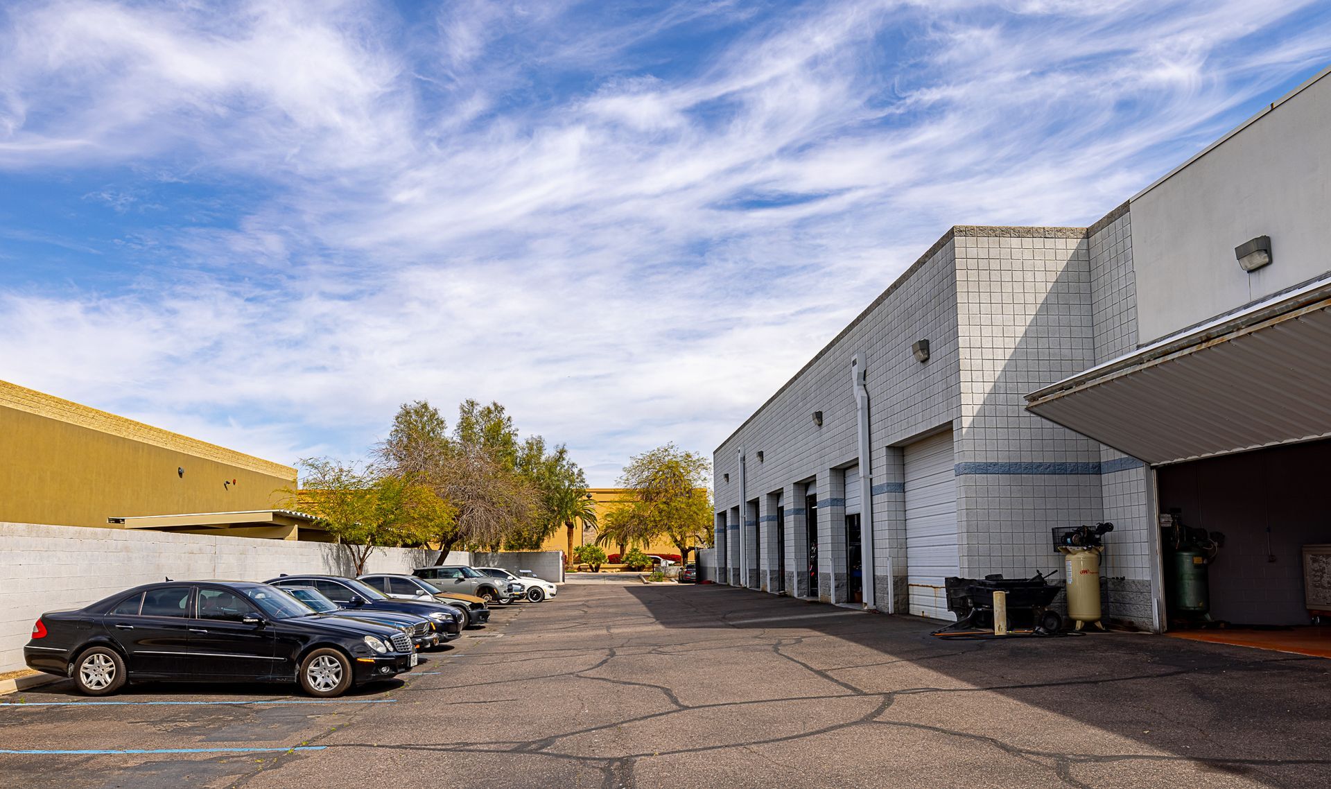 Cars parked in front of a white building with several garage bays, under a blue sky with clouds.