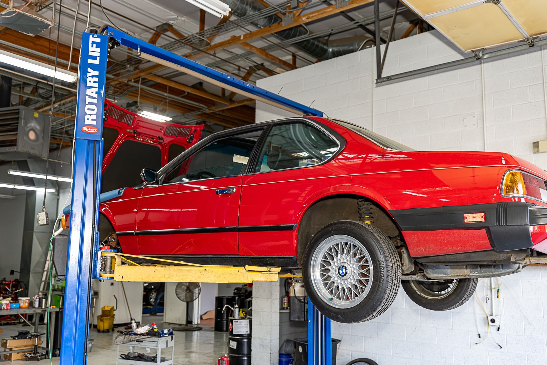Red car on a car lift in a garage, hood open, blue lift, tools and supplies visible.