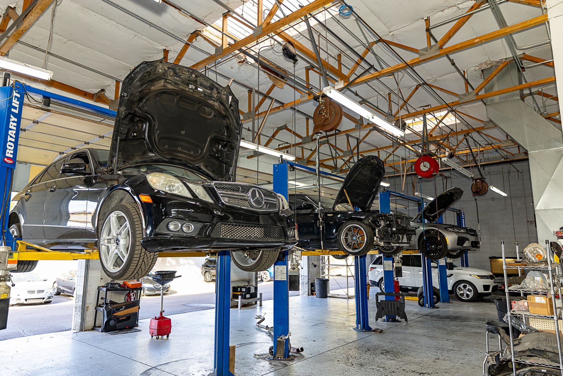 Cars on lifts in a brightly lit auto repair shop, hoods open. Workers are visible, along with tools and equipment.