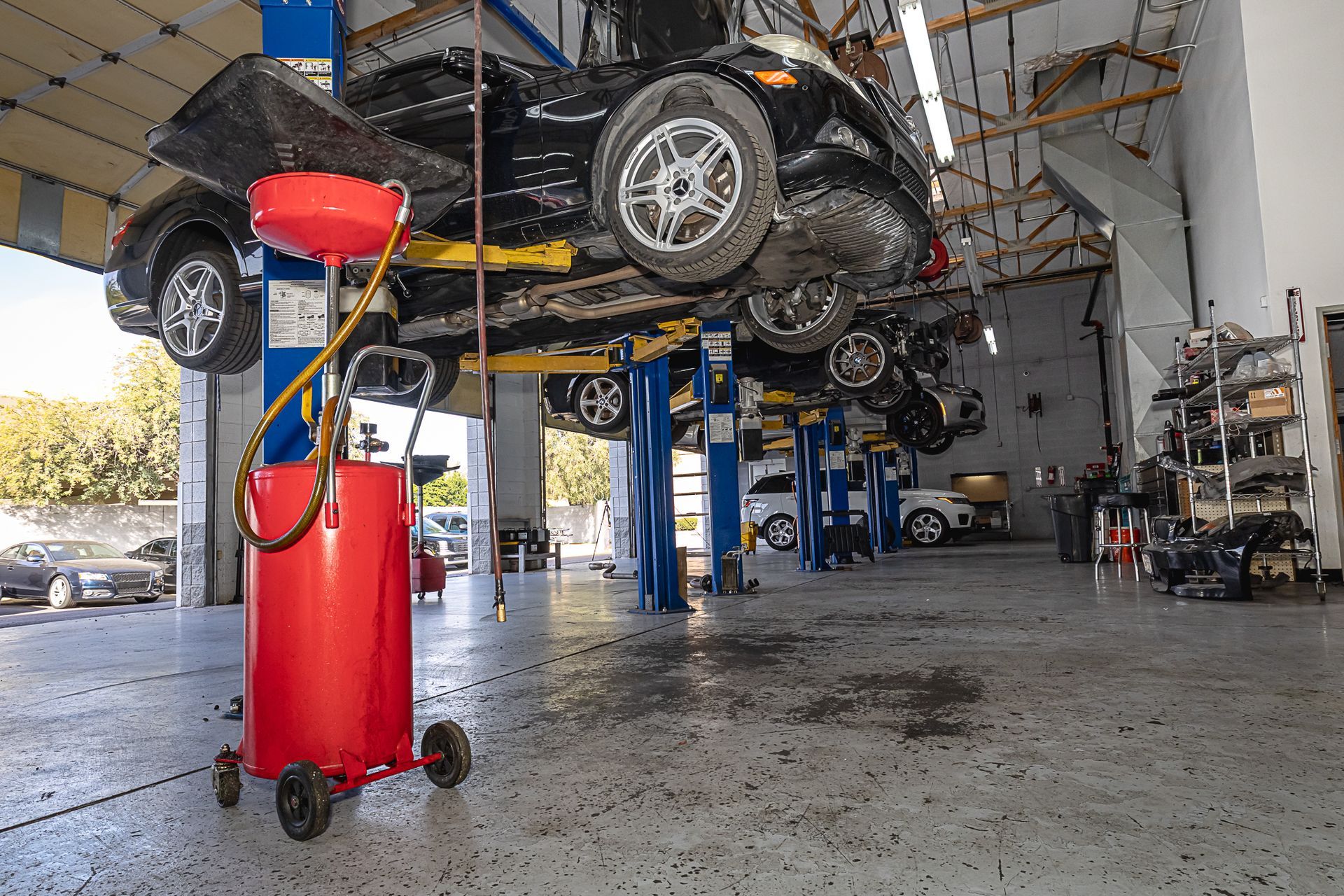 Inside a car repair shop, cars elevated on lifts. A red oil drainer is in the foreground.