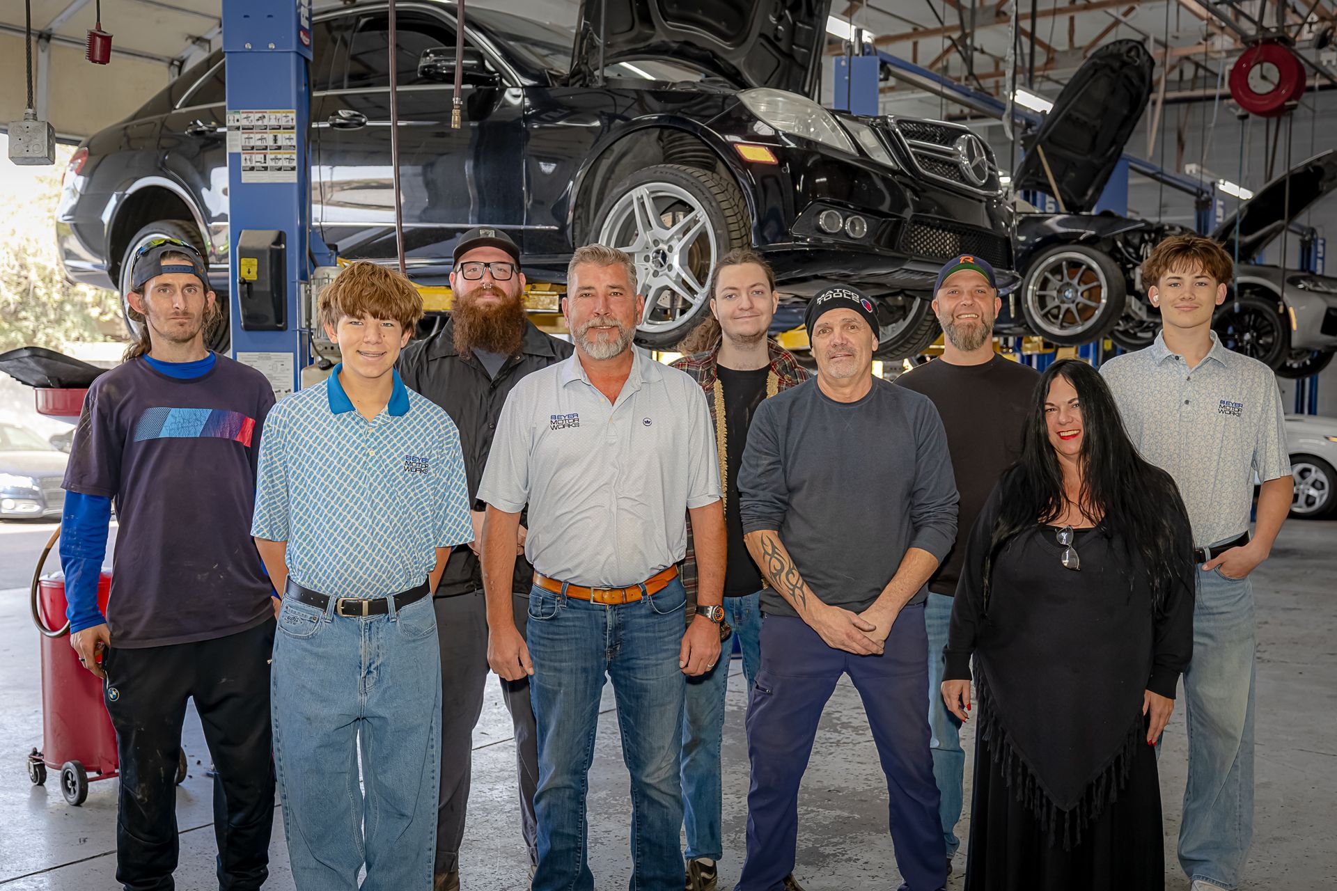 Group of people posing in a car repair shop with a car on a lift in the background.