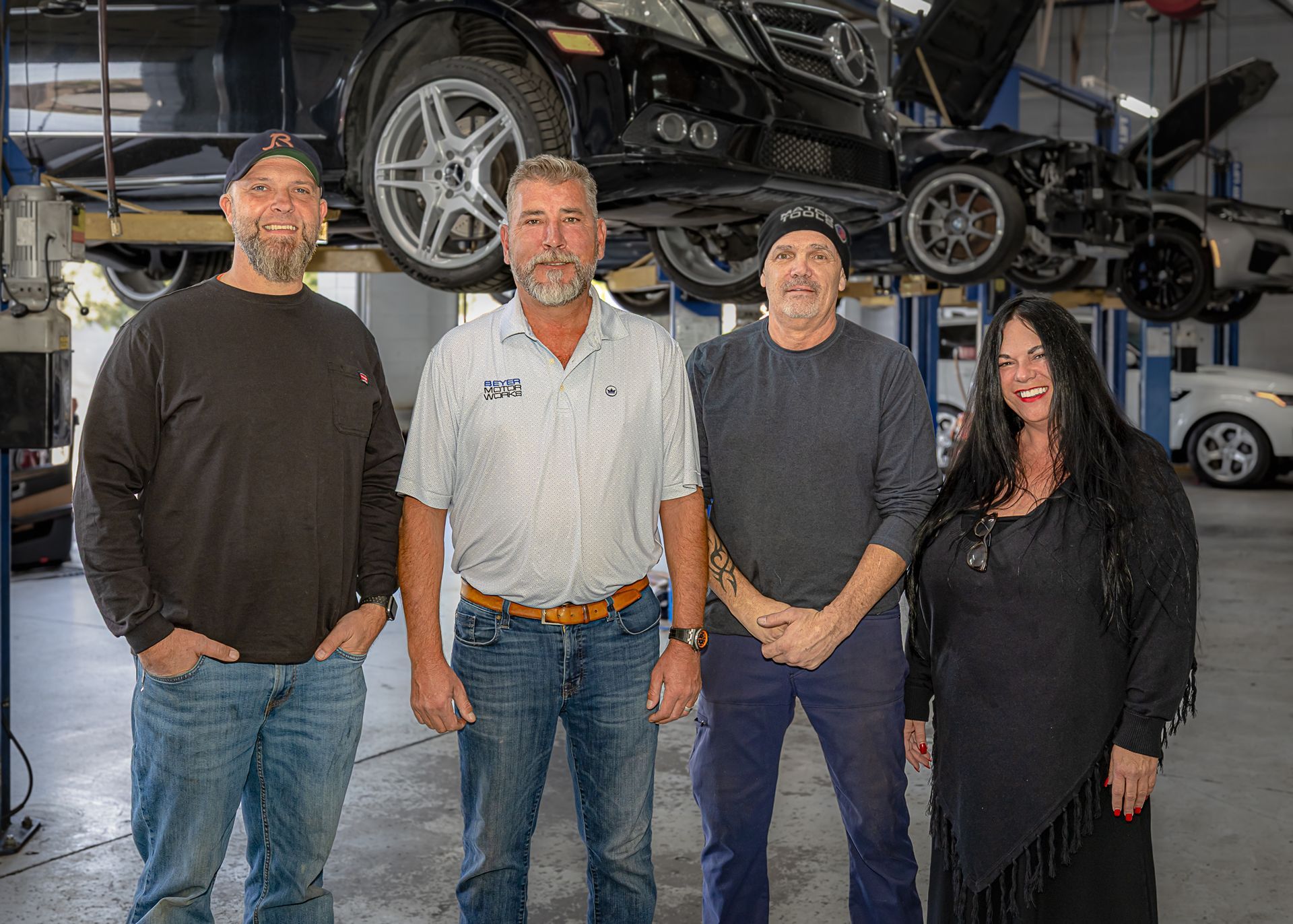 Four people in a car repair shop posing in front of vehicles on lifts.