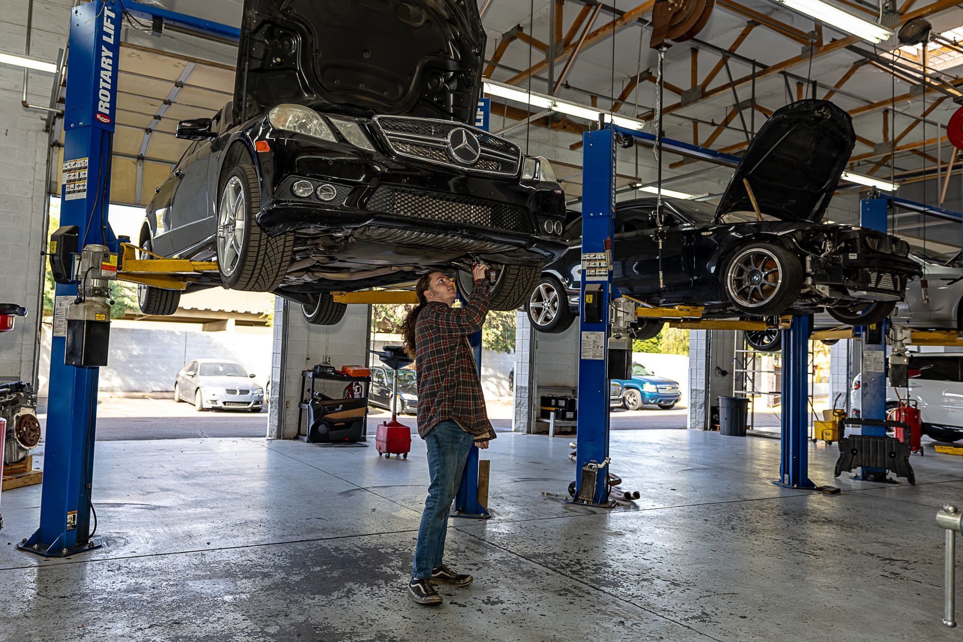 A mechanic working under a car lifted on a hoist in a garage. Another car is on a lift in the background.