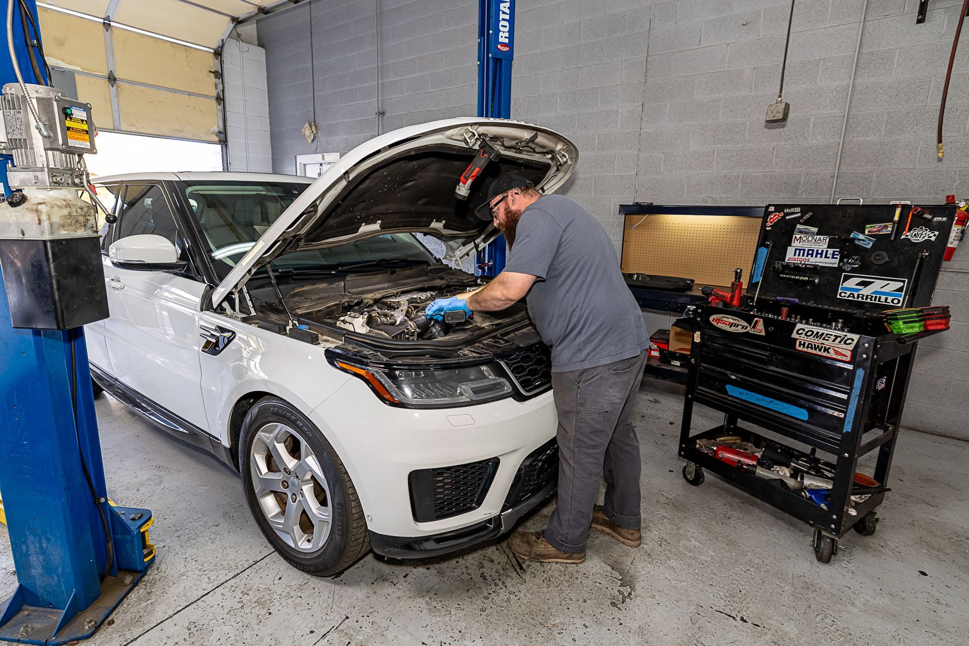 Man working on a white SUV in a garage, hood open. Tools are nearby.