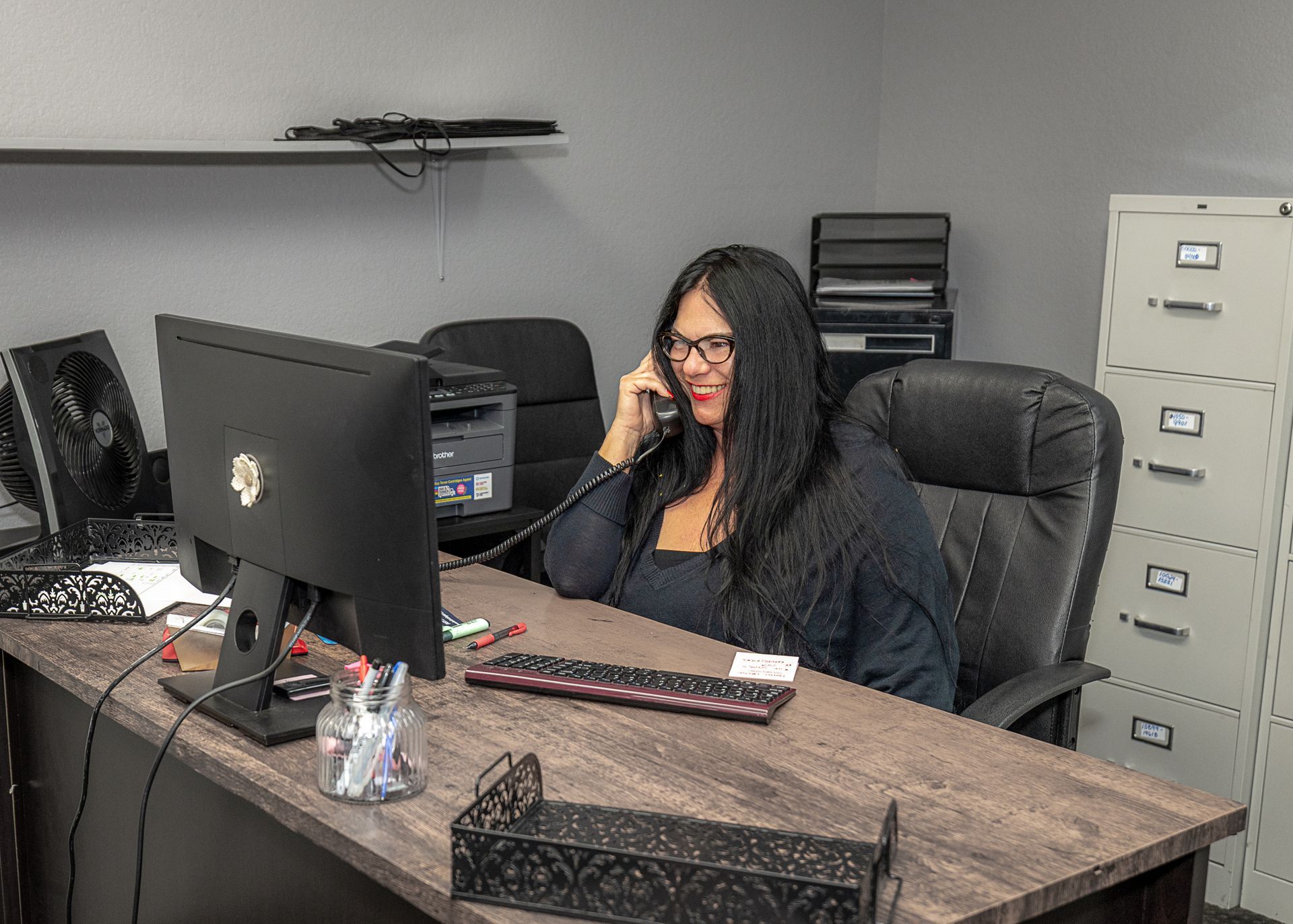 Woman in glasses talking on the phone at a desk with a computer and printer in an office.