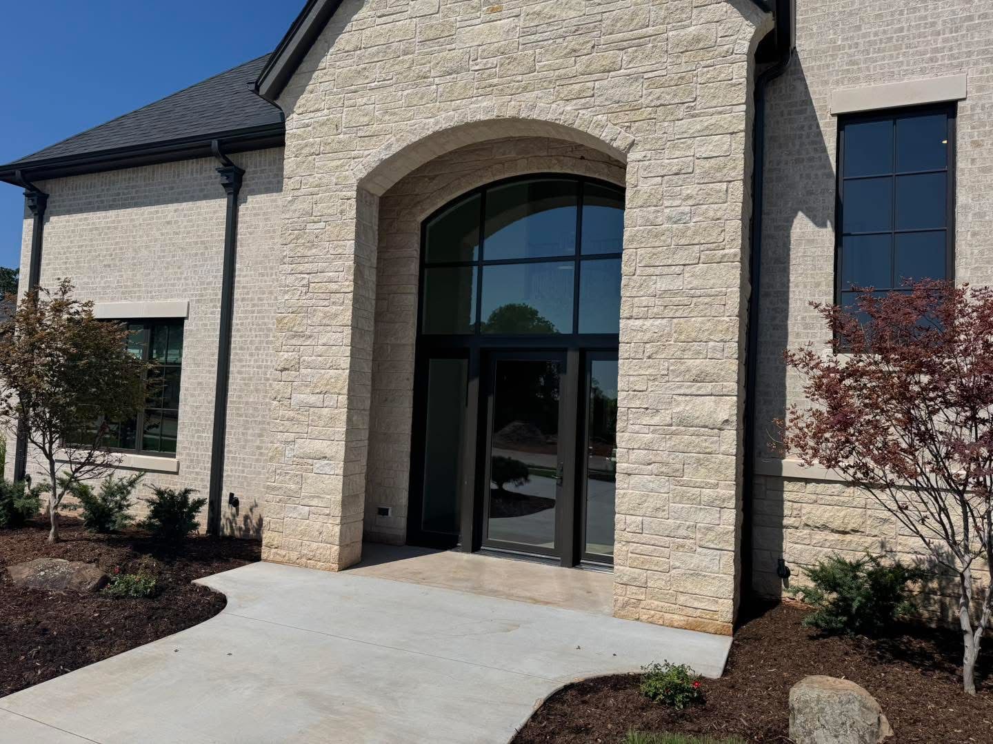 Stone building entrance with arched doorway, black-framed glass door, and windows. Dark roof, gray walkway.