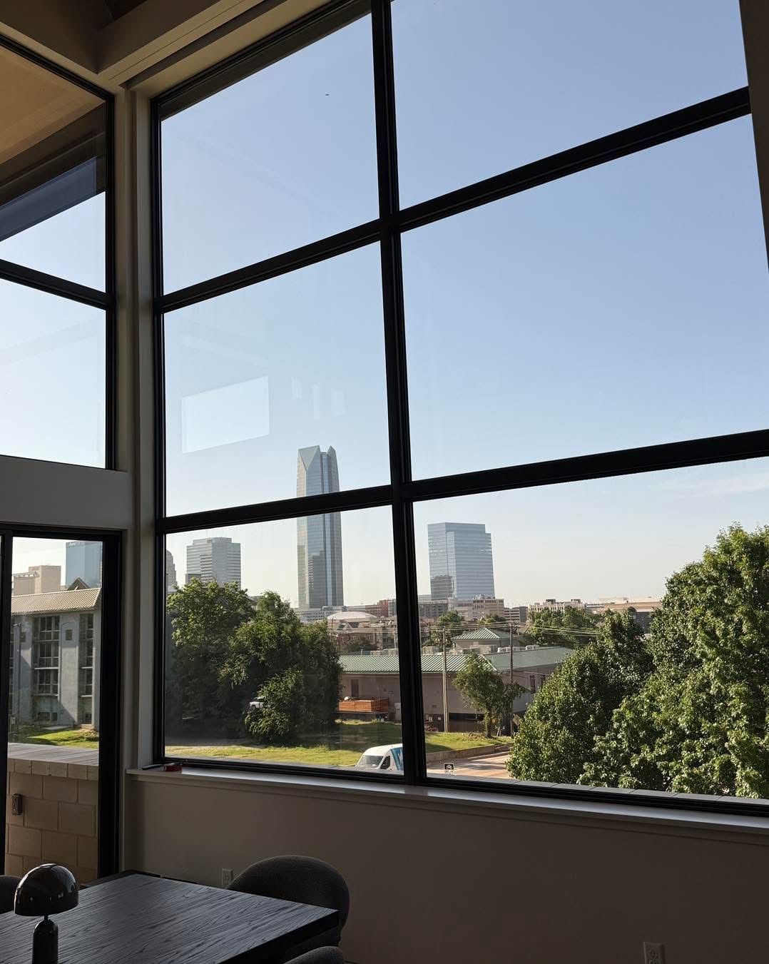 View through large windows: city skyline with tall buildings, trees, and a sunny sky. Interior table and chairs in foreground.