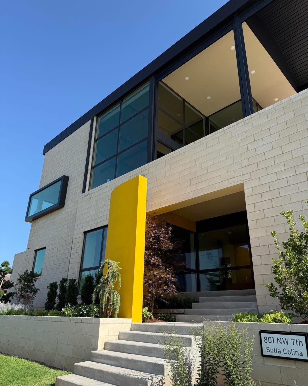 Modern two-story house with light brick exterior, large windows, and a bright yellow accent wall, leading to the entrance.