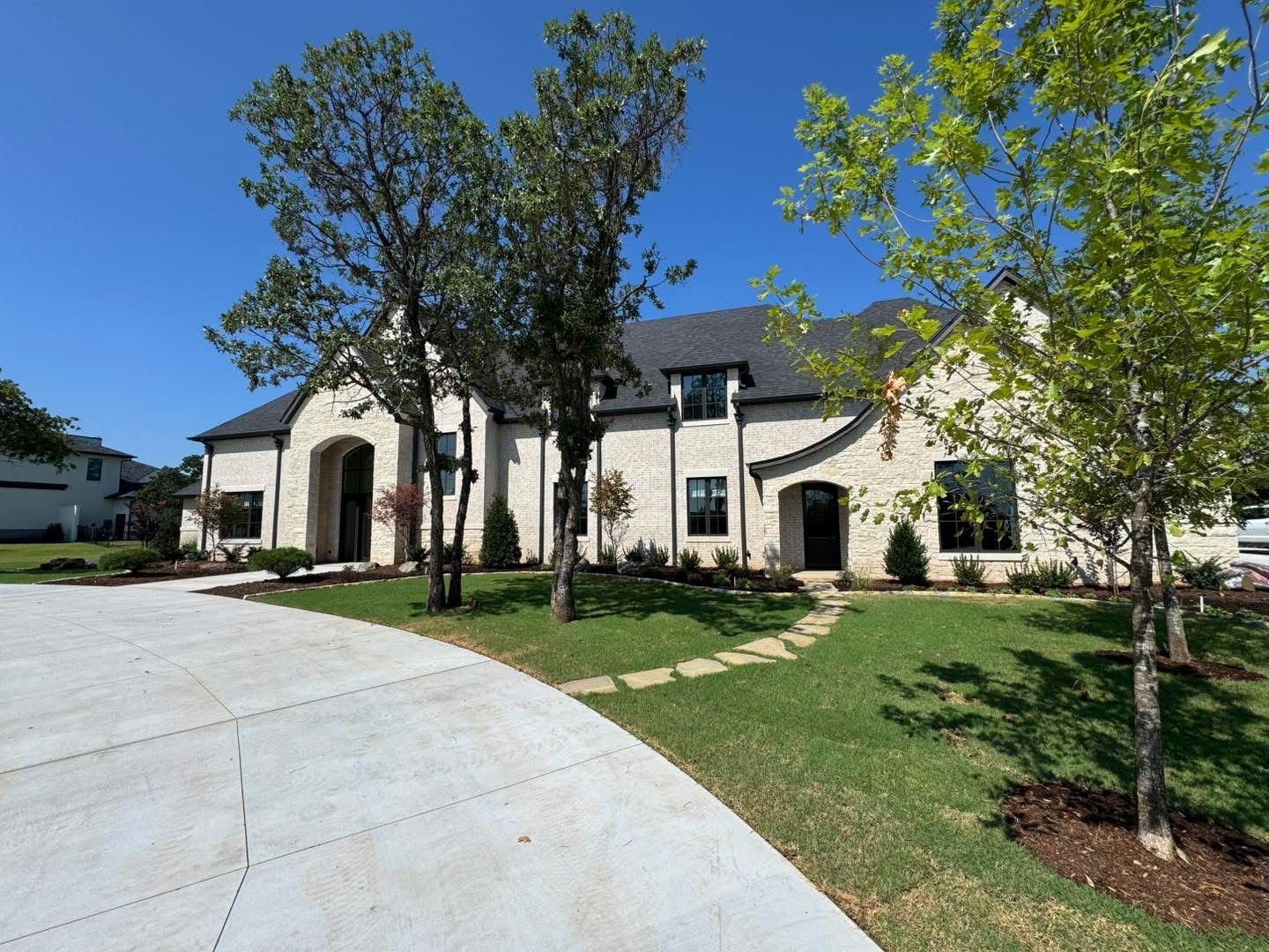 Two-story stone house with arched entry and dark roof, set on green lawn, with trees in front and curved concrete driveway.