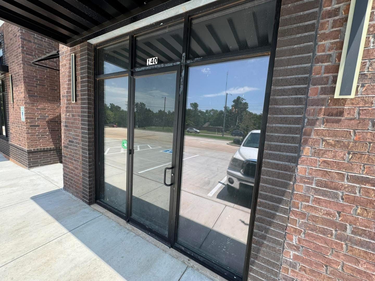 Exterior of a commercial building with glass doors reflecting the parking lot. Brick facade, awning, sunny day.