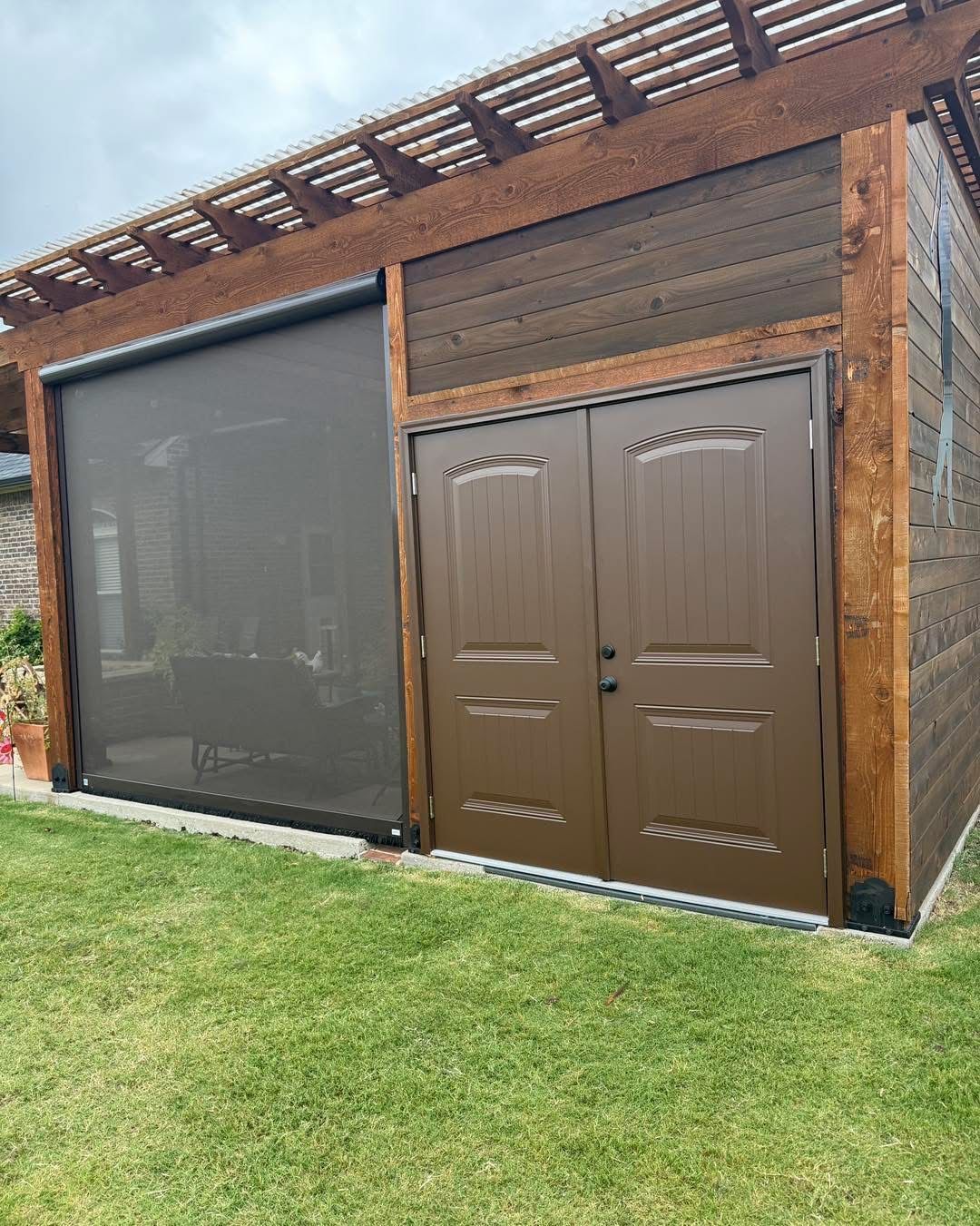 Wooden structure with roll-down shade, brown double doors, and slatted roof; on green grass.