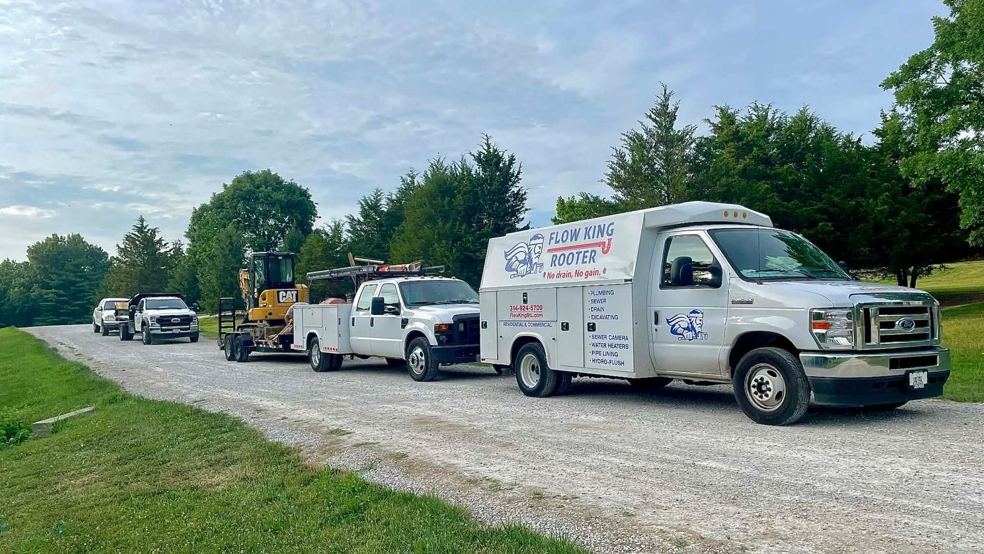 Line of white service vehicles with equipment on a gravel road, trees in the background, blue sky.
