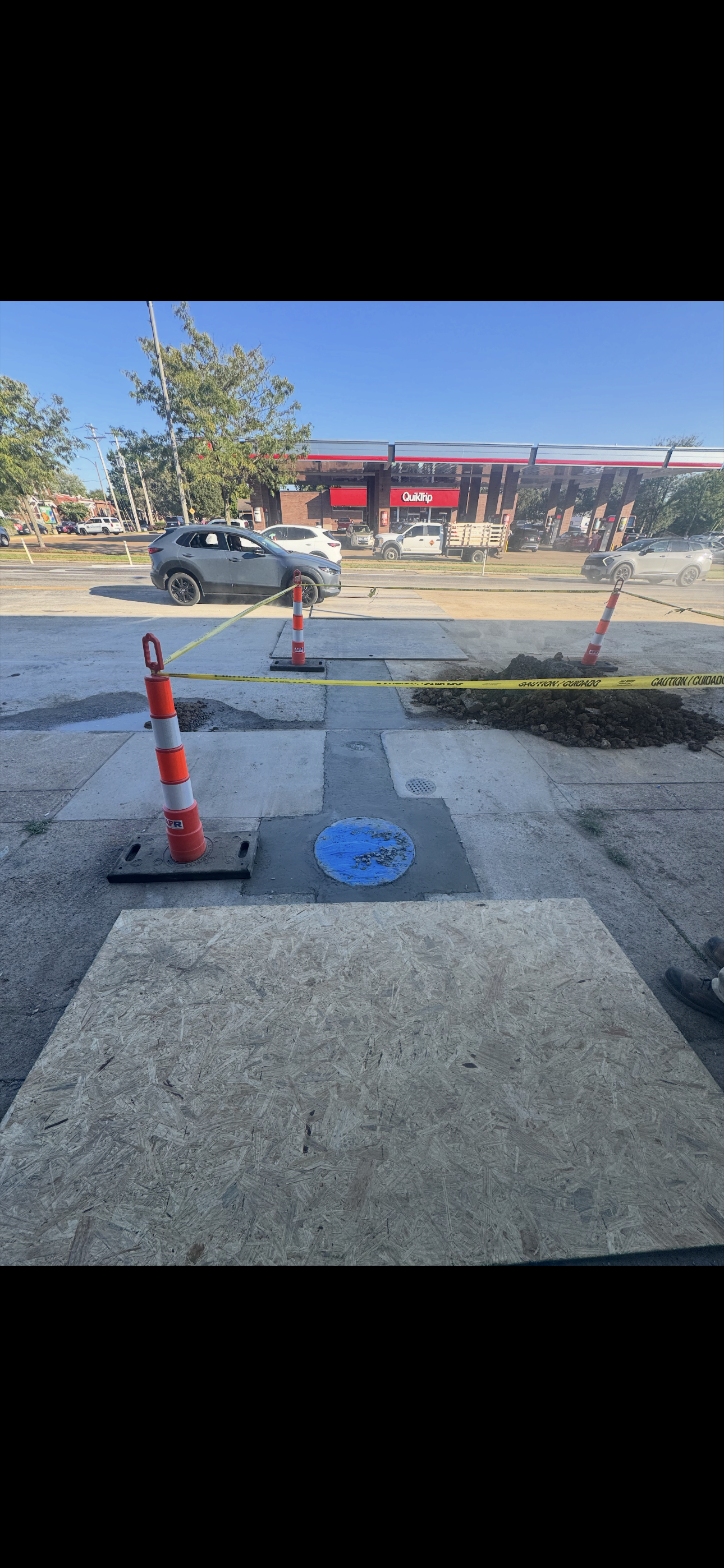 Asphalt road with orange cones and blue circle around a manhole cover, with a building in the background.