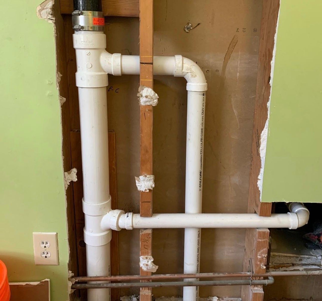 White PVC pipes in a wall cavity, connected to a drain and vent, with exposed studs and drywall.