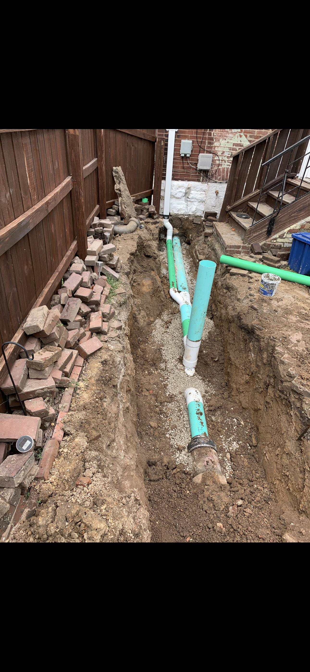 A trench with green and blue pipes, bricks, and a wooden fence.