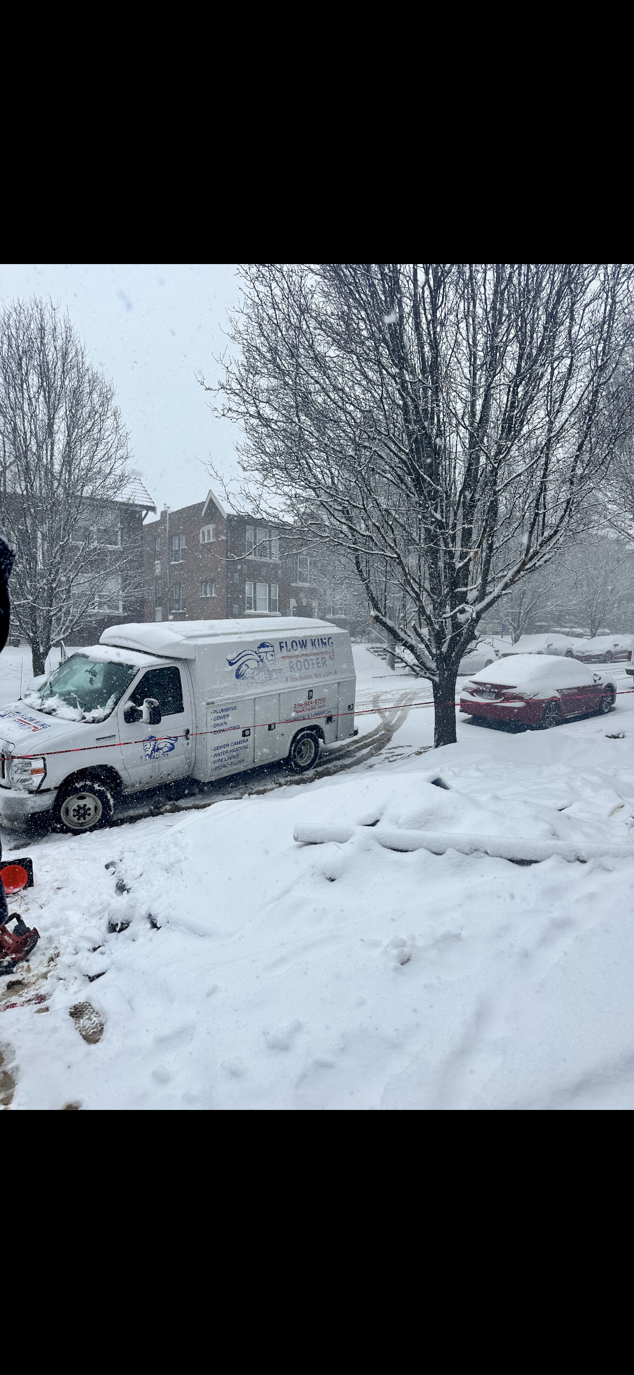 Snow-covered white van and red car on a snowy street, a tree on the right, and more buildings in the background.