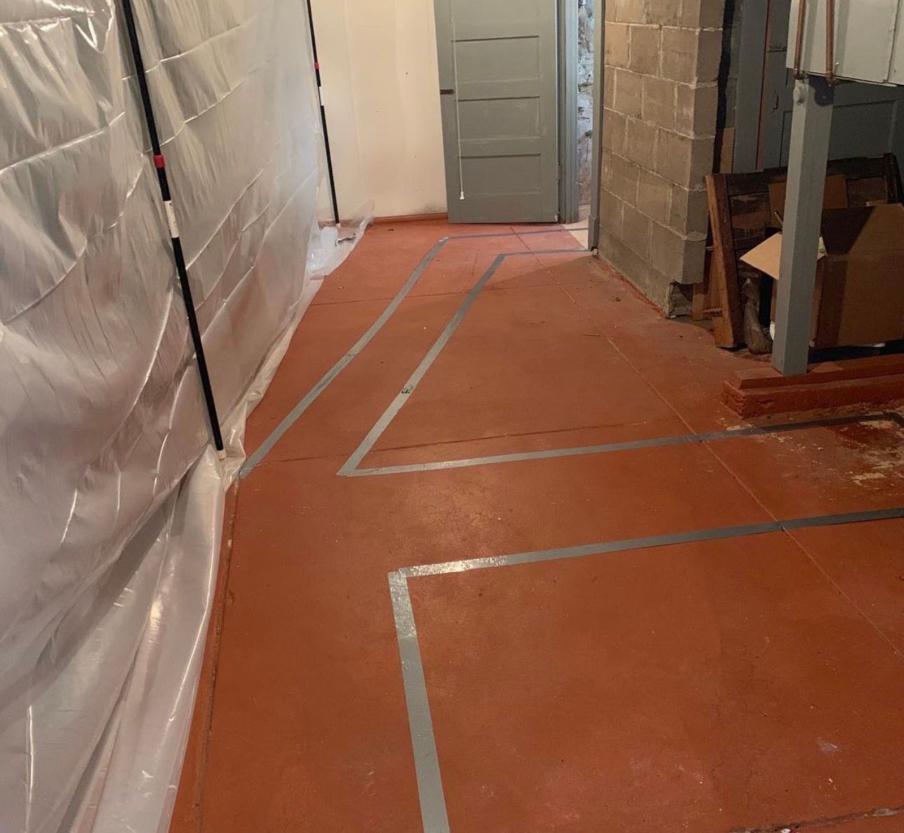 Red floor with silver tape, leading toward a gray door in a basement. White plastic on the left wall.