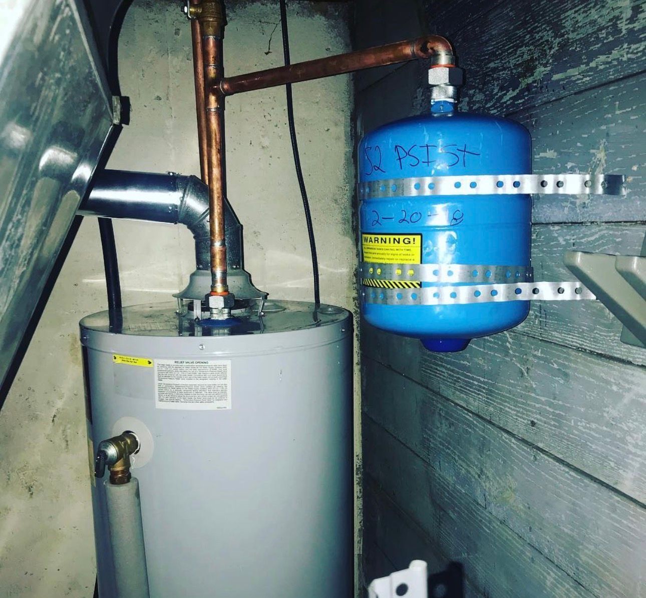 Water heater with blue expansion tank and copper pipes attached to a wall in a utility room.