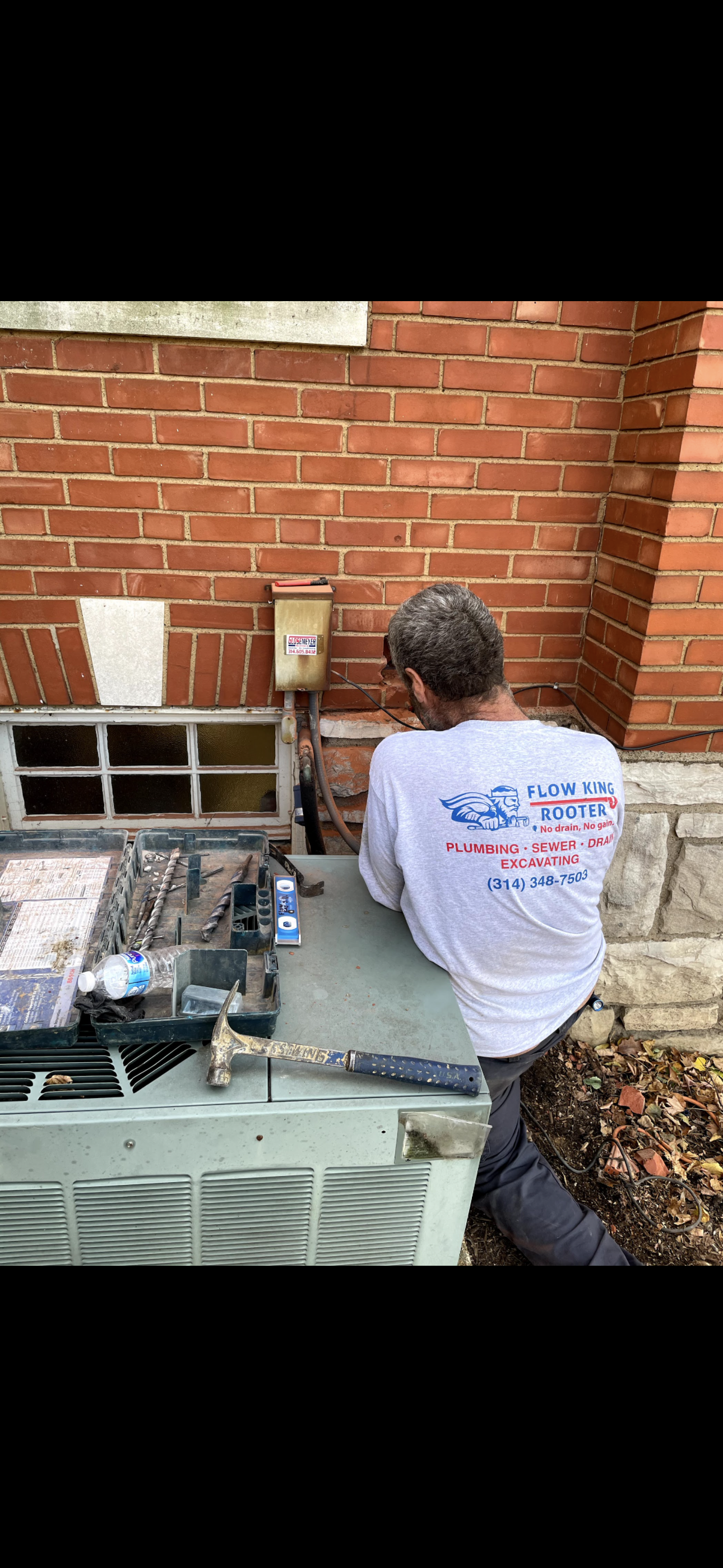 HVAC technician working on an air conditioning unit near a brick wall and a foundation.