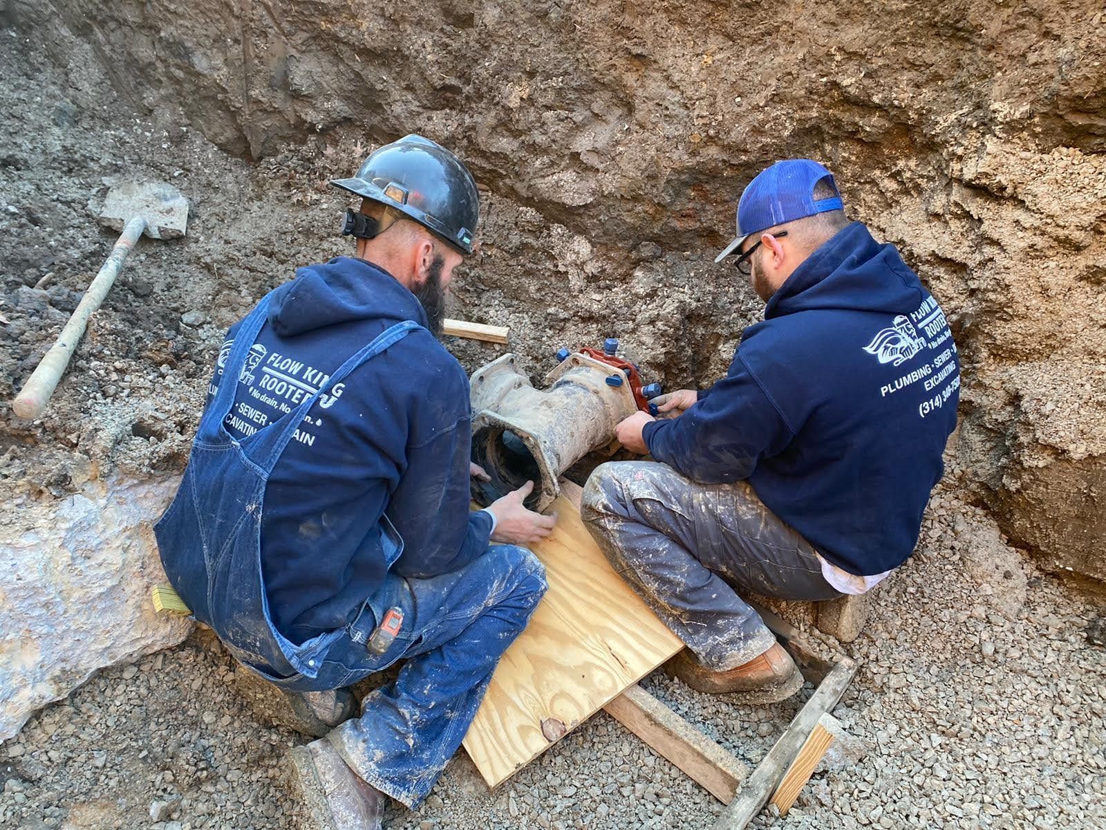 Two plumbers repairing pipes in a trench. One in overalls, the other in a hoodie.