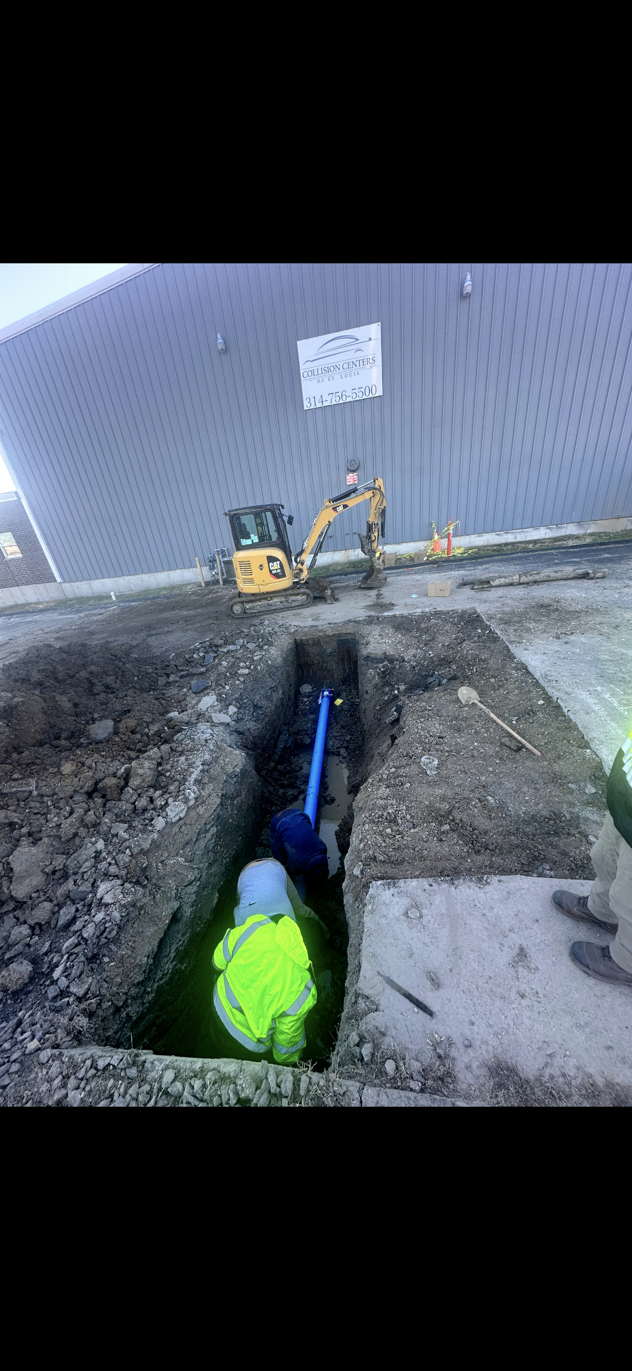 A worker in neon yellow vest in a trench, with machinery and building in the background.
