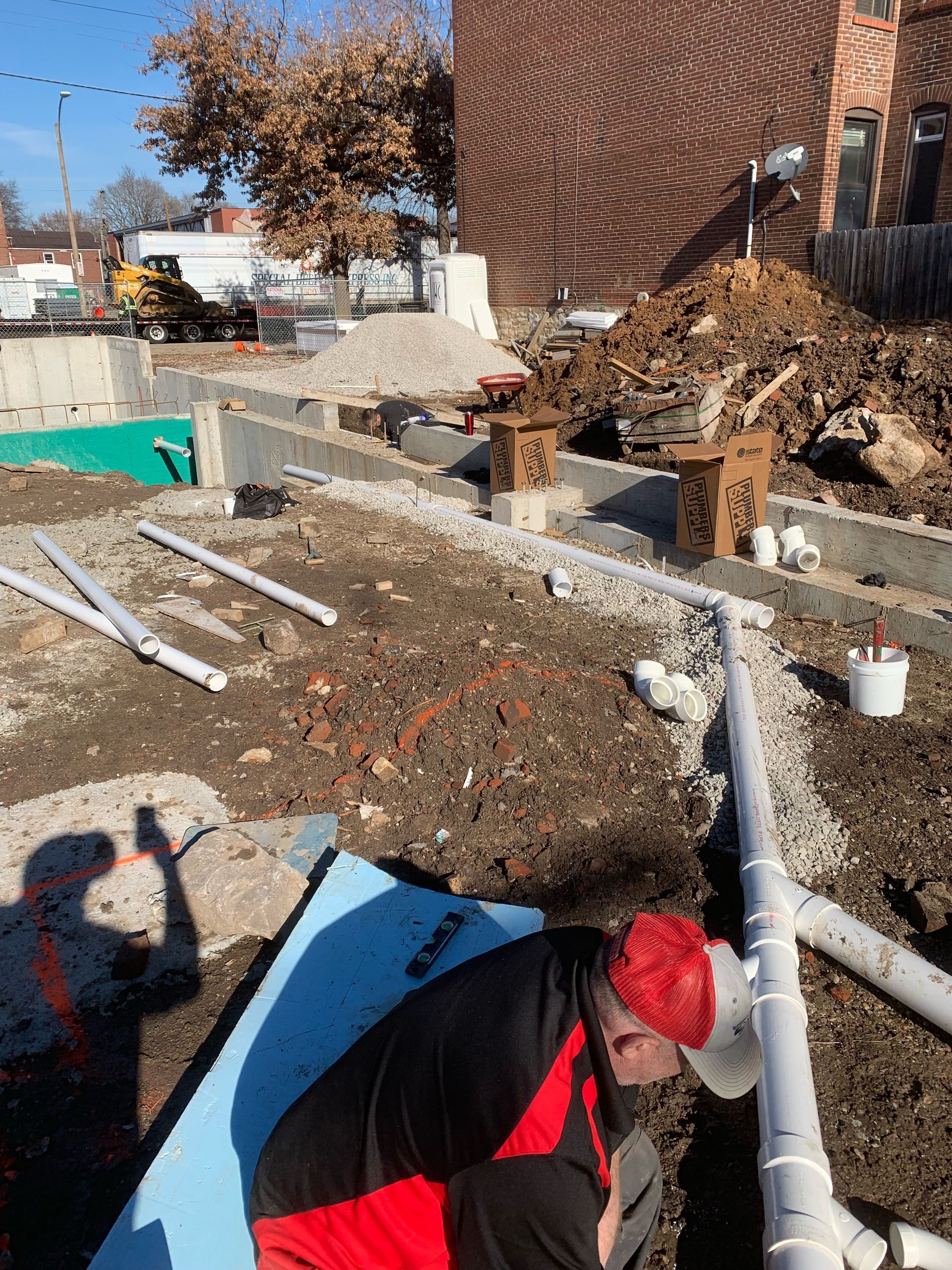 Construction worker in red and black outfit installs pipes at a building site with dirt piles and foundation.