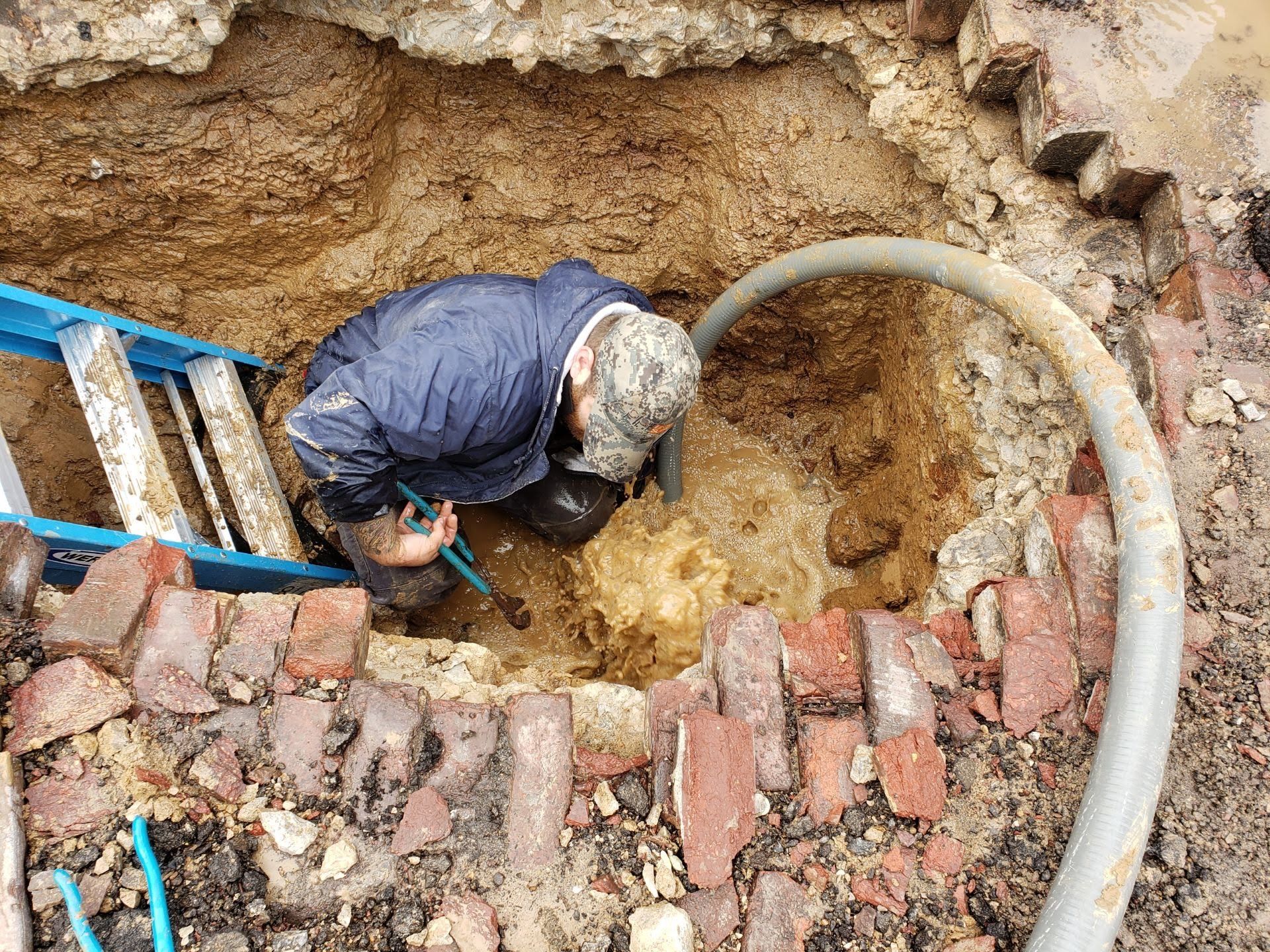 A person repairs a pipe in a muddy, excavated hole with a ladder and hose.