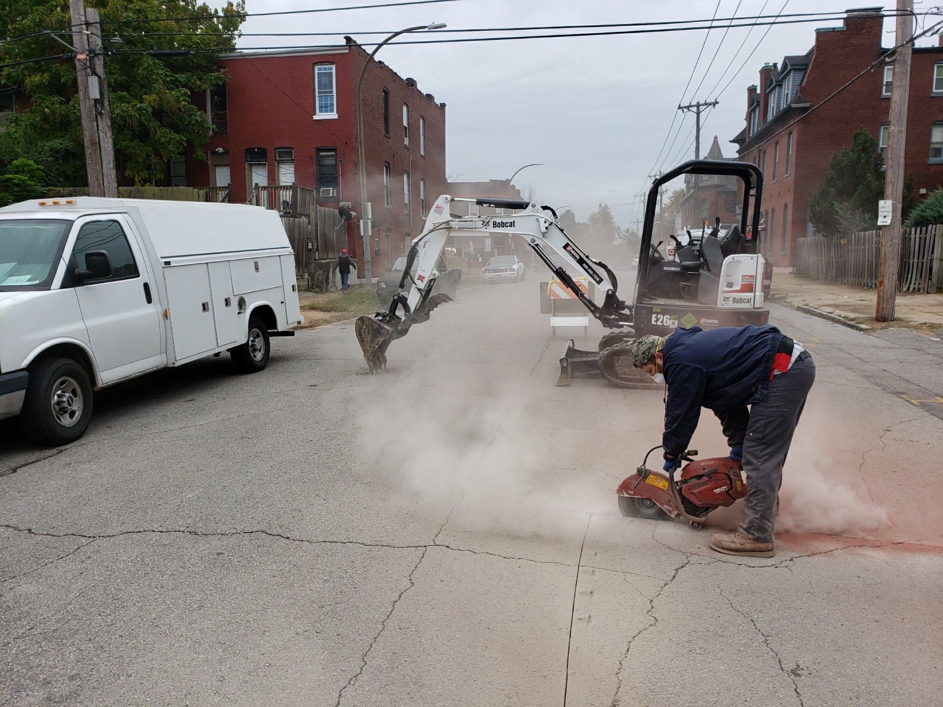 Man using saw to cut road, excavator behind. White van parked on street with brick buildings.