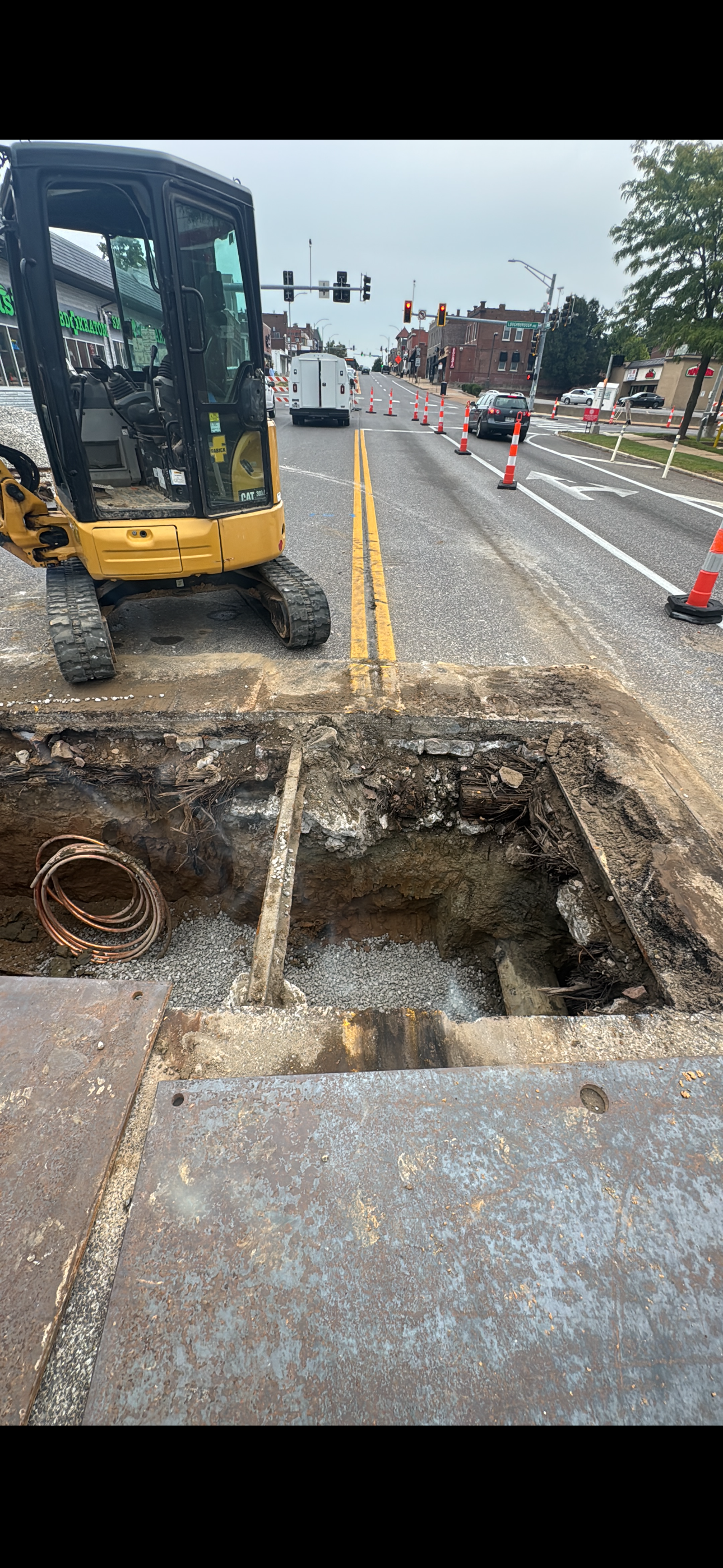 A small excavator sits near an open excavation on a road, orange cones guide traffic, and buildings line the street.