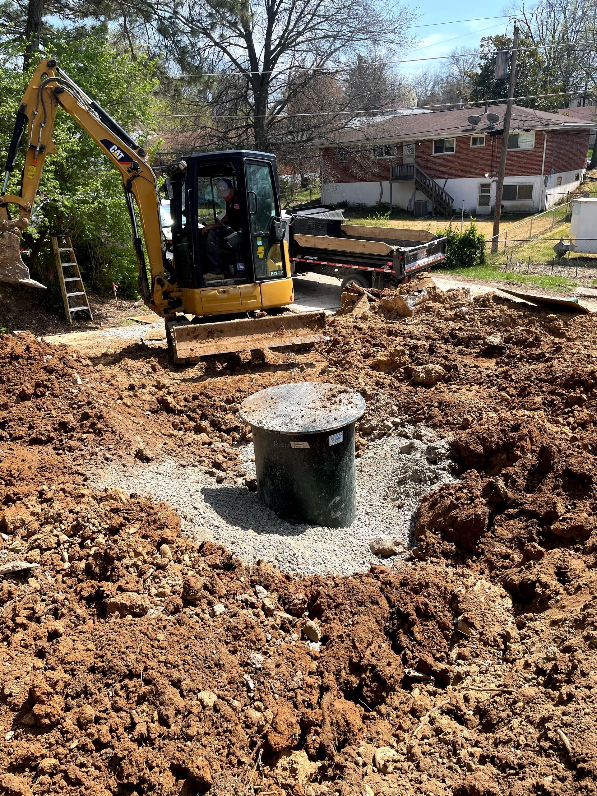 Excavator digging around a septic tank. Brown dirt surrounds the tank, with houses in the background.