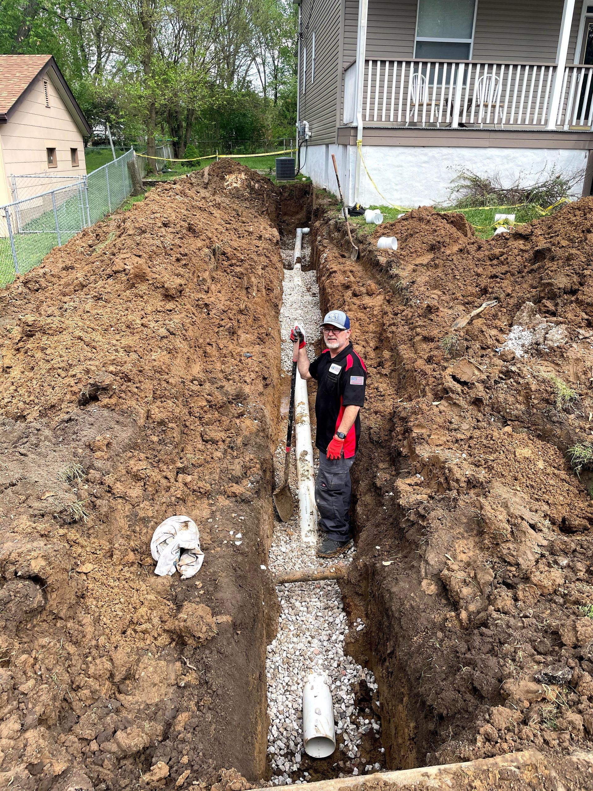Man in a trench installing pipes; dirt and gravel surround. House in the background.