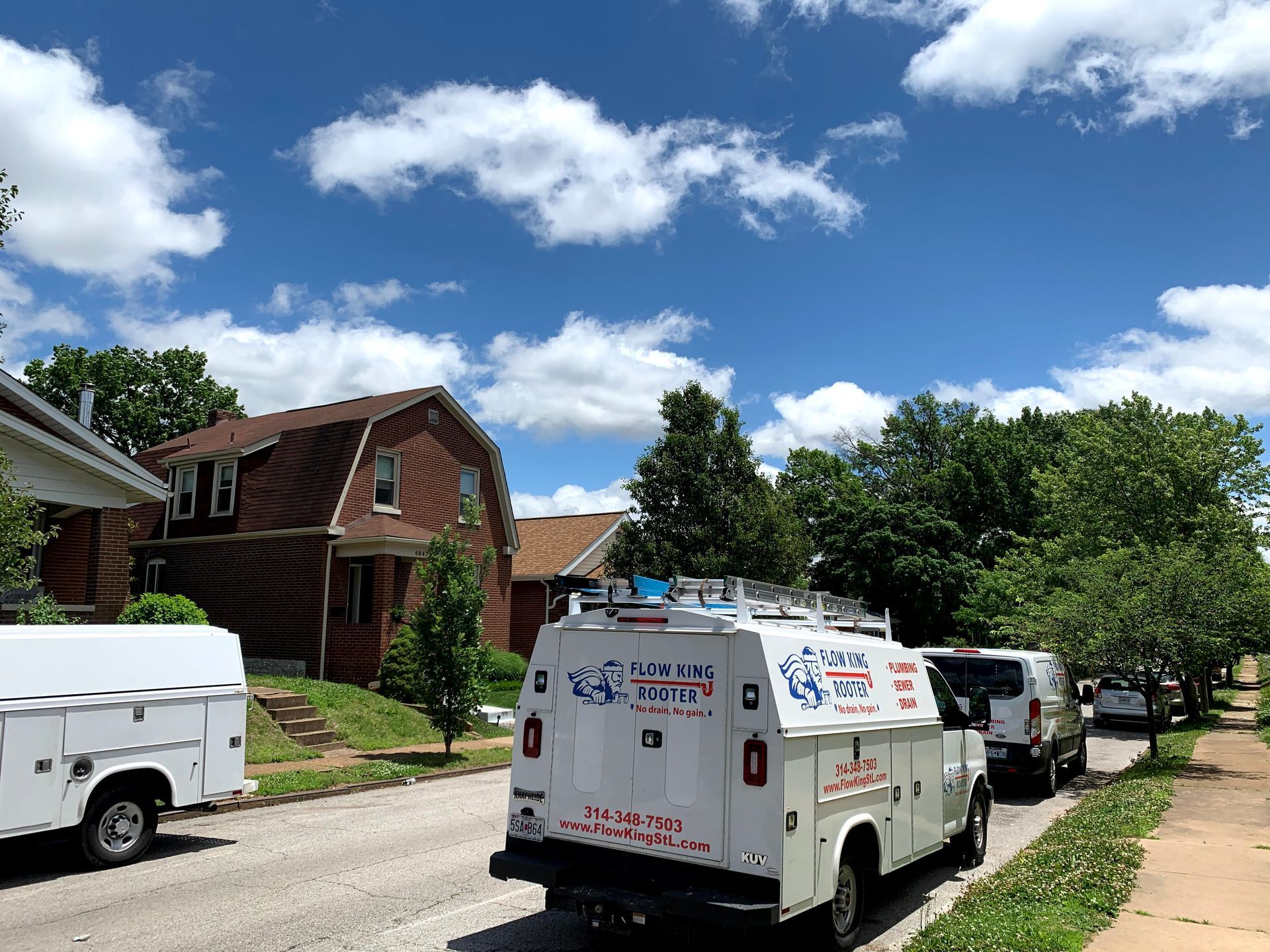 Service vans parked on a residential street under a blue sky with fluffy clouds. Brown house visible.