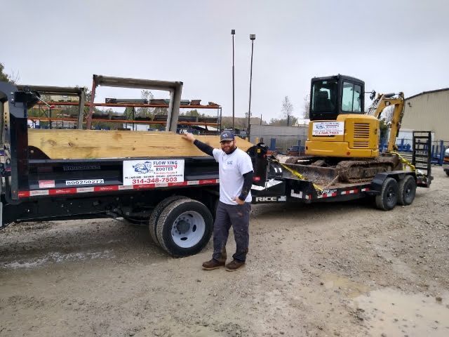 Man stands by a dump truck and a trailer with an excavator; outdoor, overcast day.