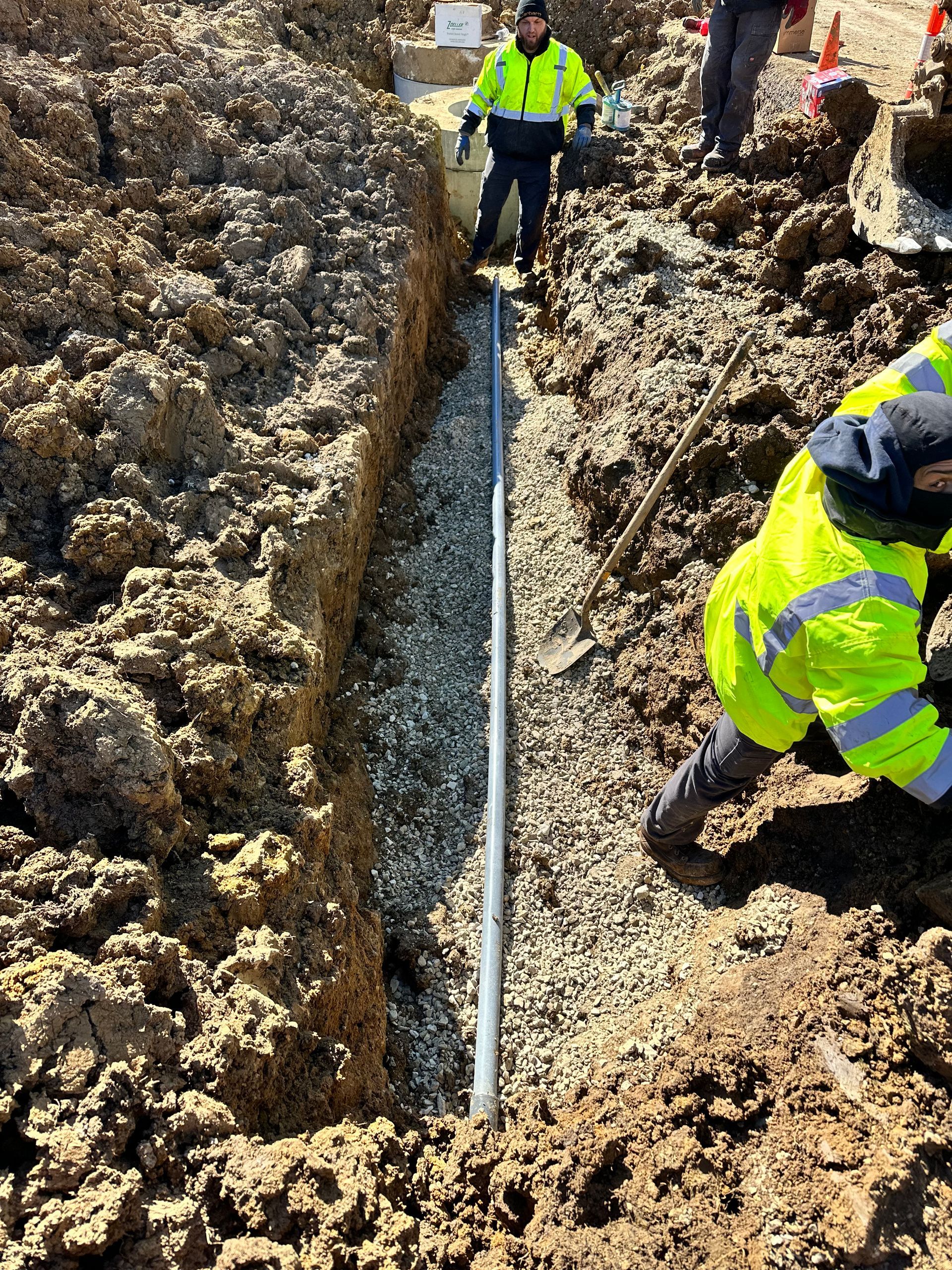 Workers in reflective vests install pipe in a trench filled with gravel.