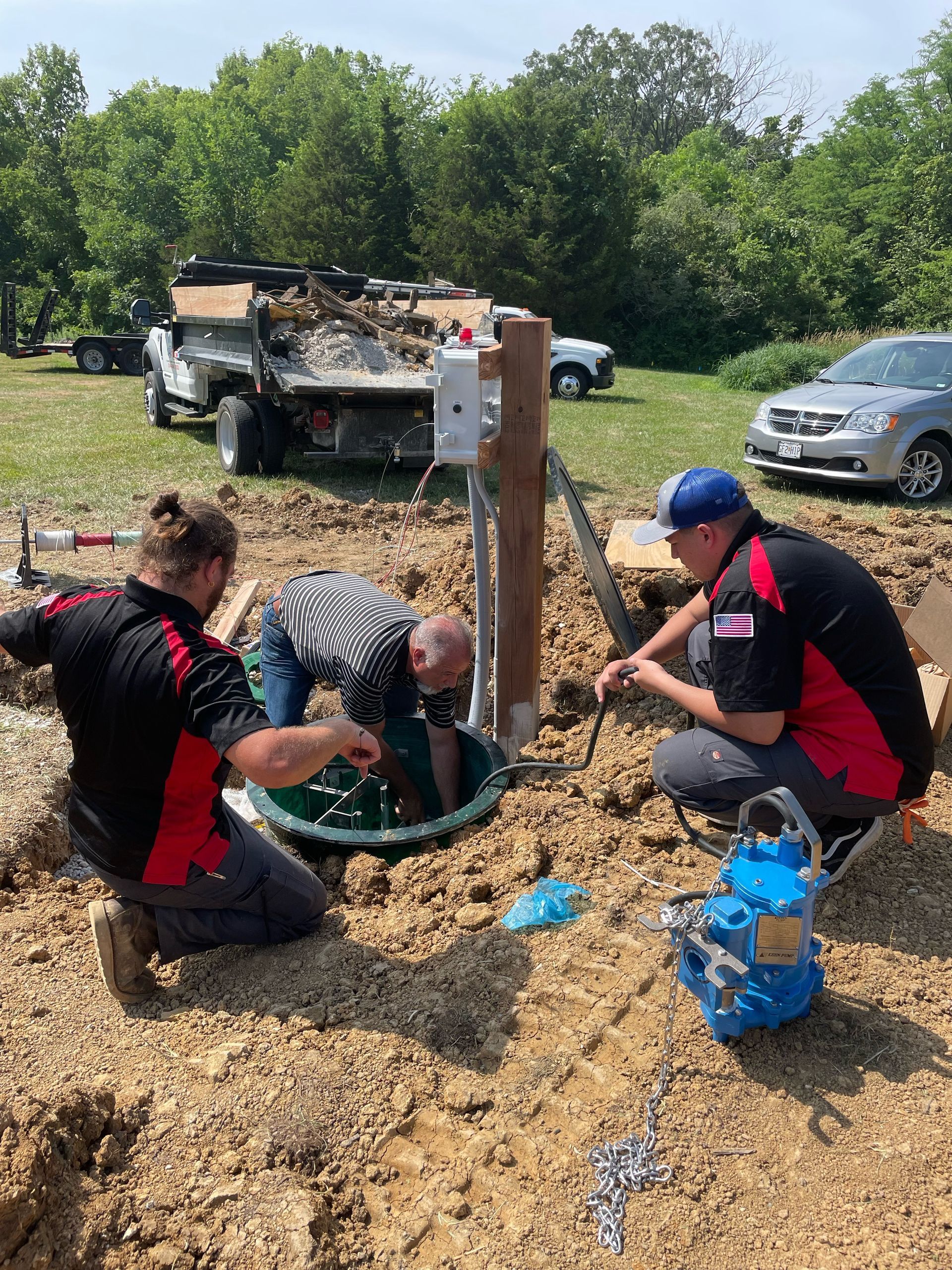 Three workers installing equipment near a well, outdoors on a sunny day.