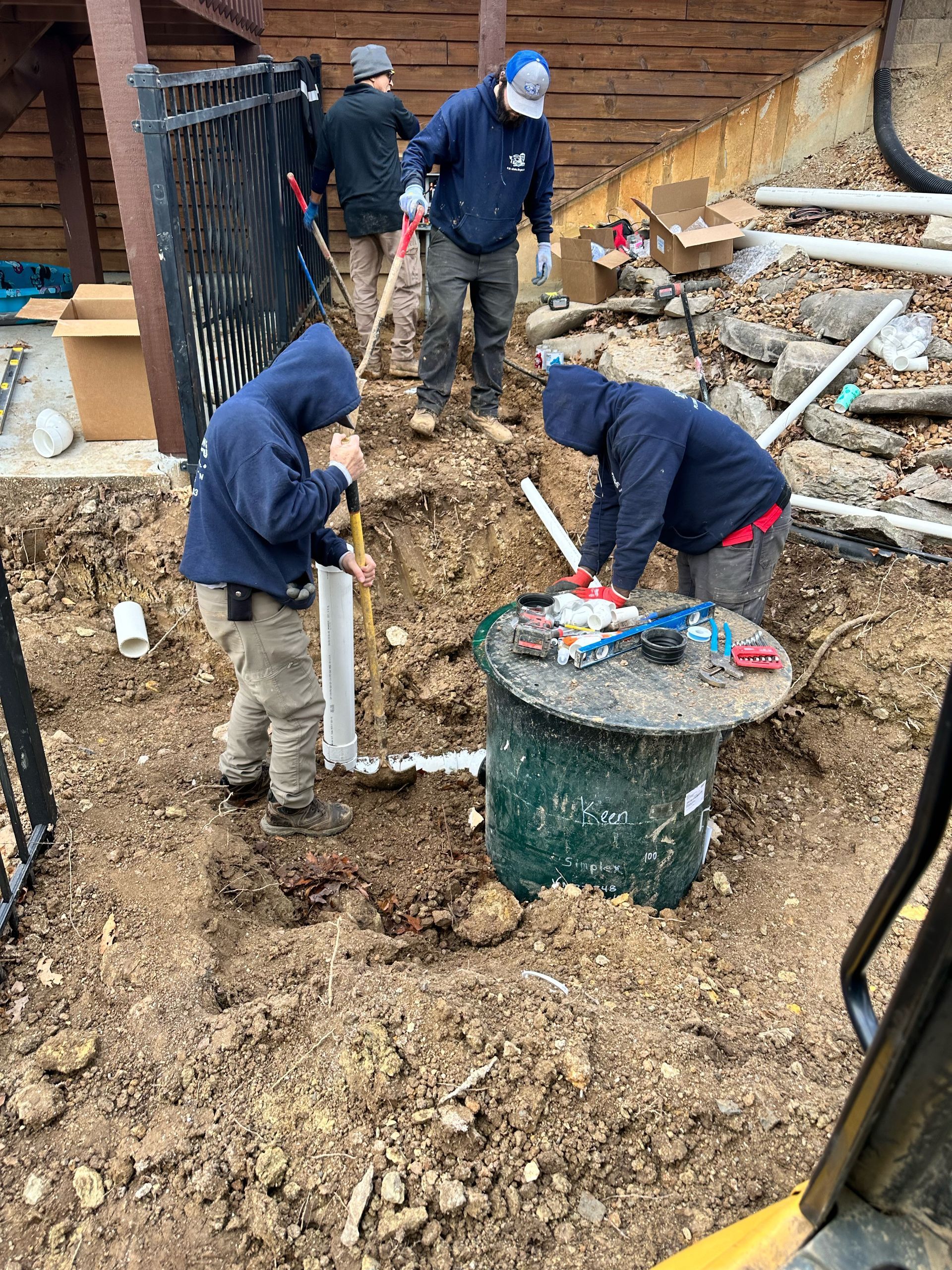Workers installing plumbing in an excavated area near a building; they are using tools and a septic tank.