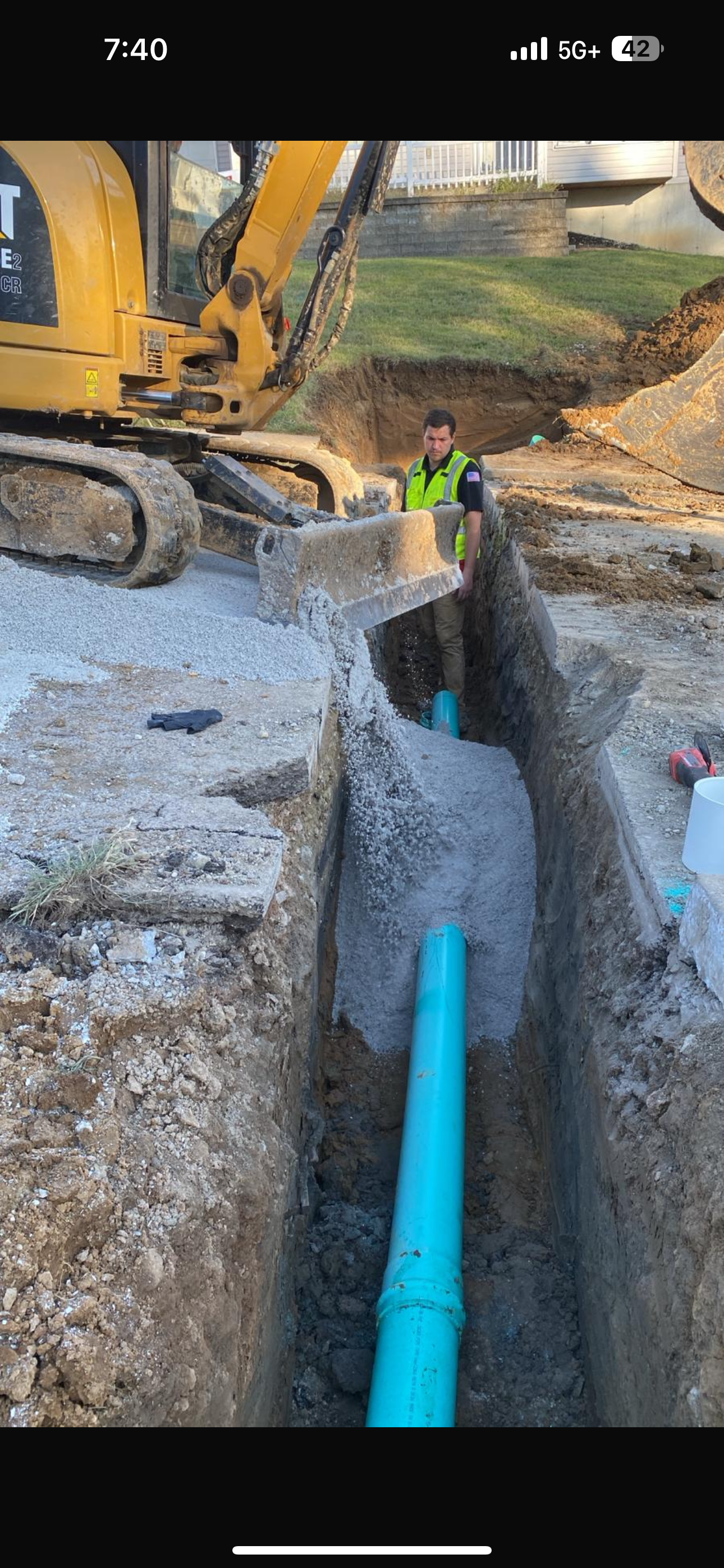 Construction site trench with blue pipe, gravel, and yellow excavator in progress. A worker in a vest is present.