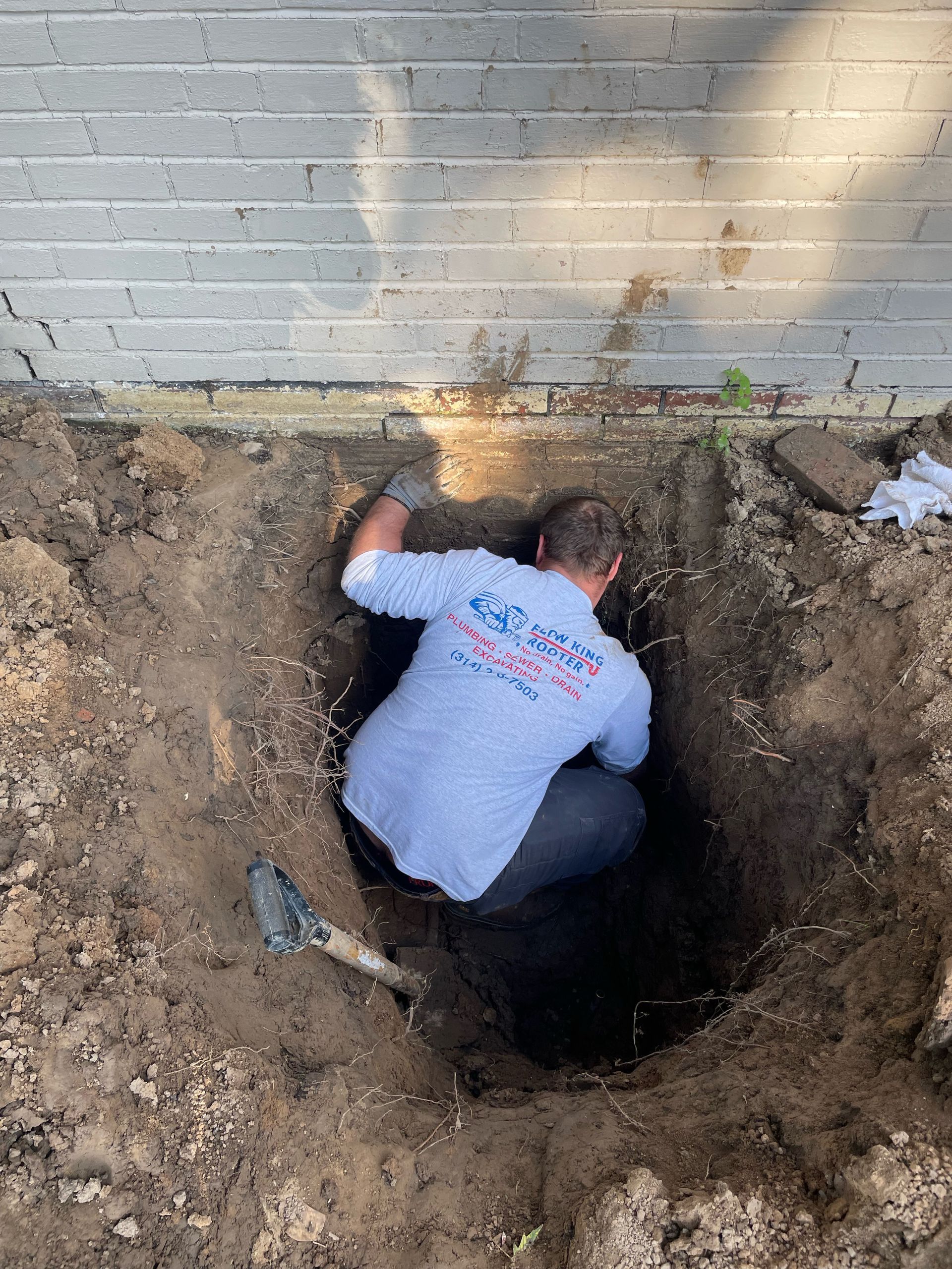 Man in a hole near a wall, wearing a white shirt with blue lettering, using tools to work.
