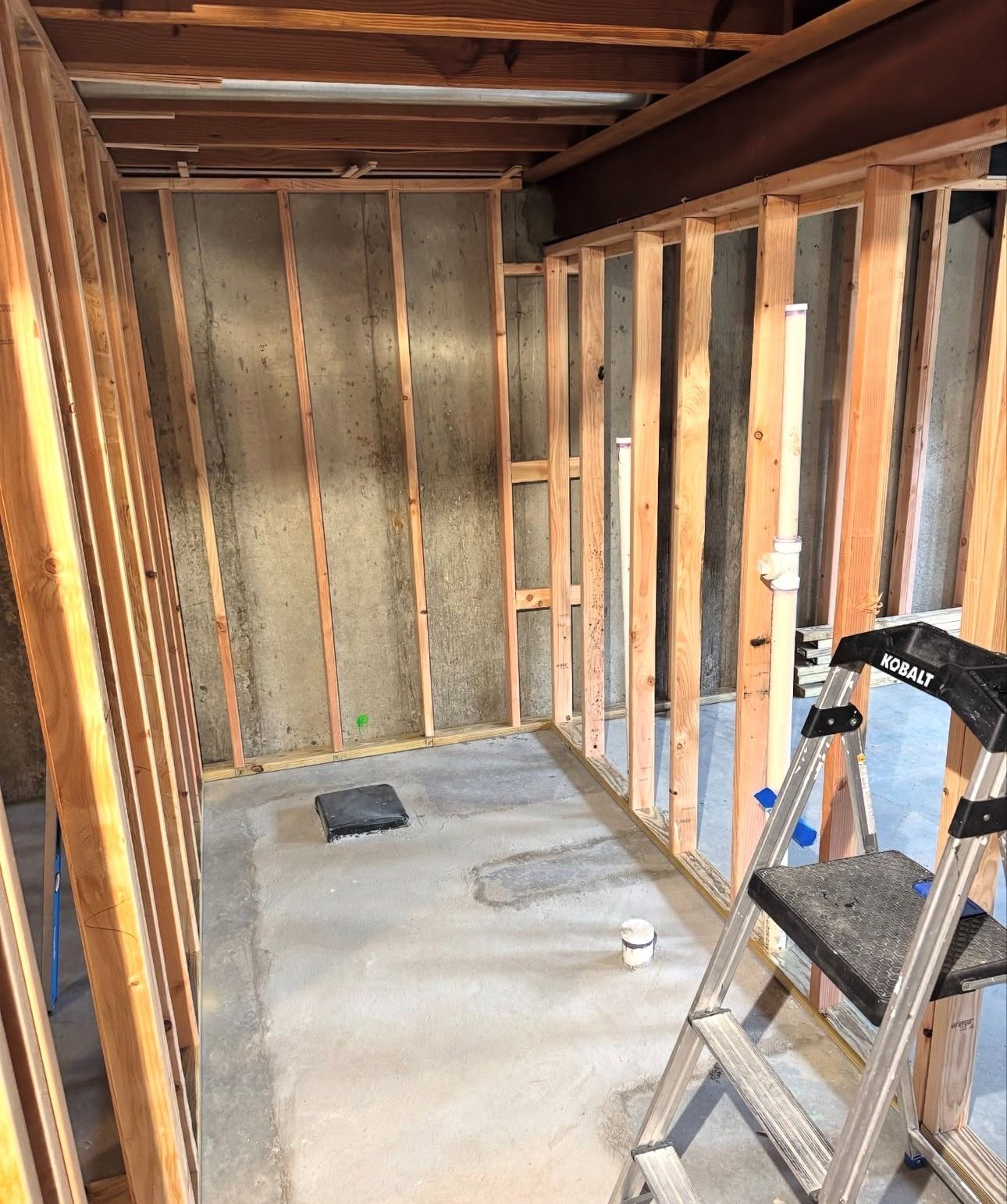 Framed interior walls with exposed concrete and plumbing; a step ladder is present.