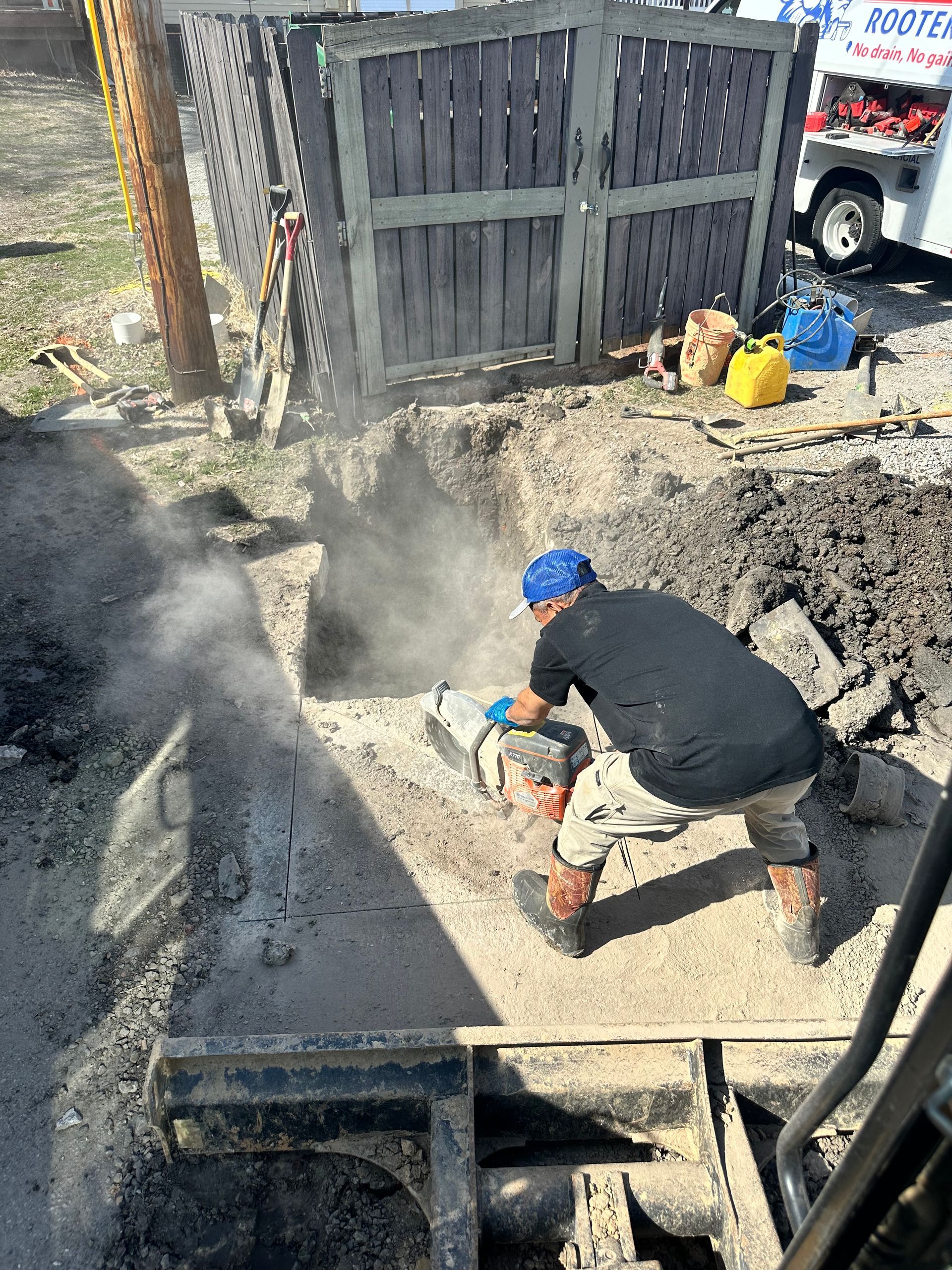A person cutting concrete in a trench; dust is present. Behind them is a fence, a truck, and supplies.