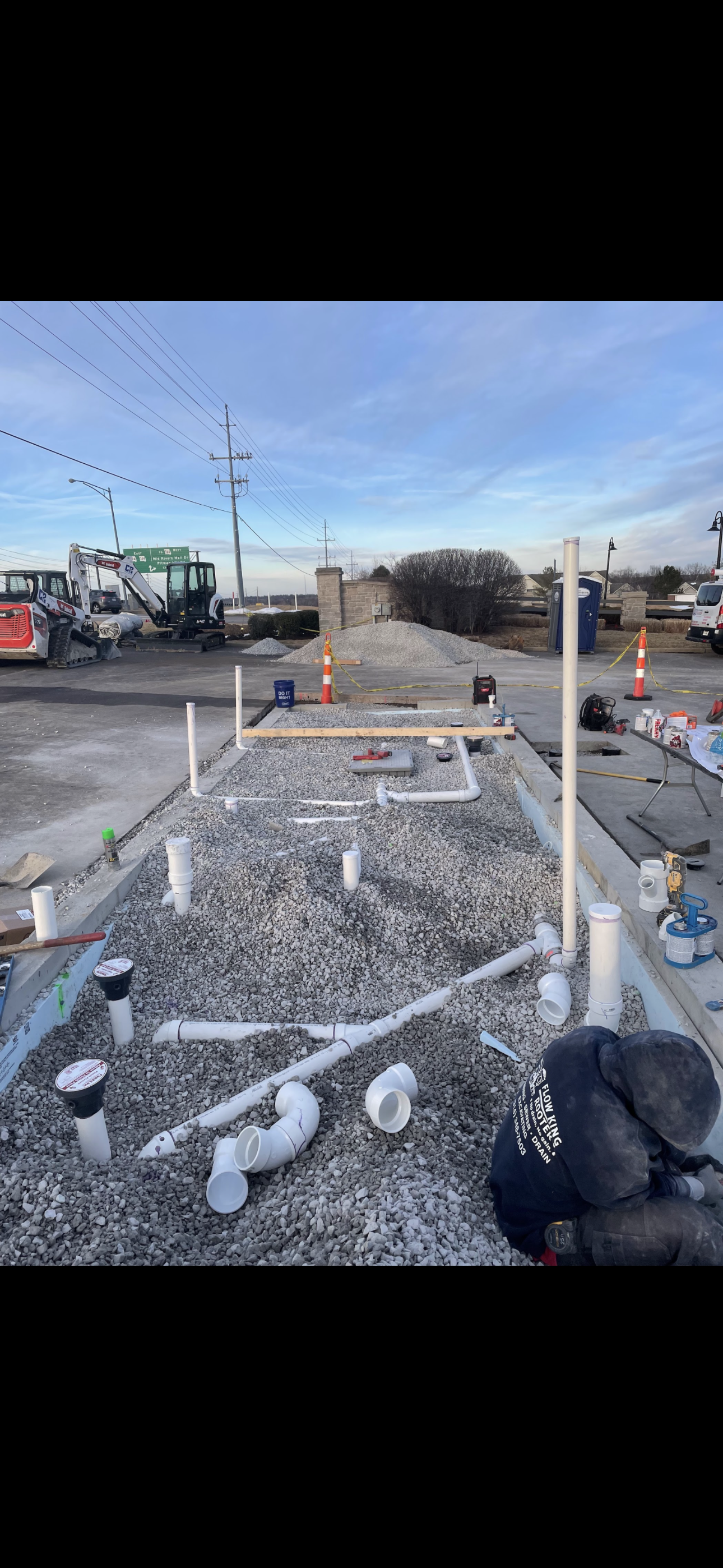 Construction site with plumbing installation in progress. Pipes, gravel, and a worker are visible.