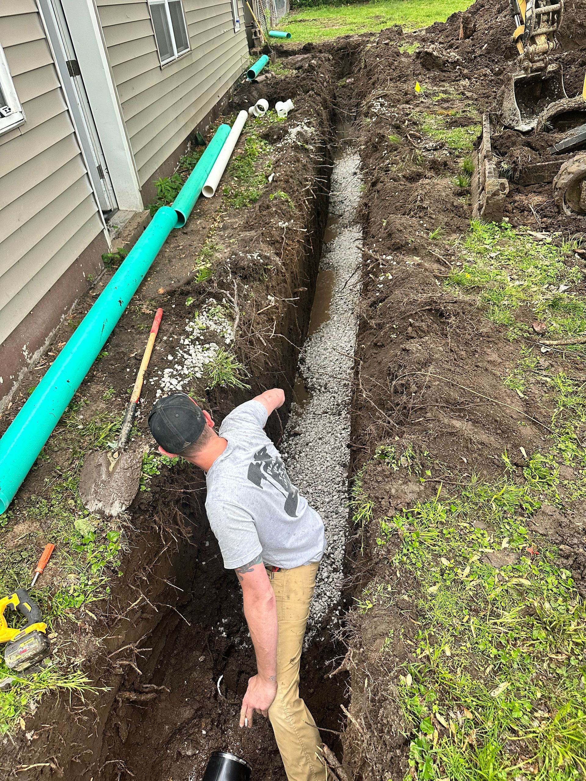 Man installing drainage pipe in trench filled with gravel next to a house.